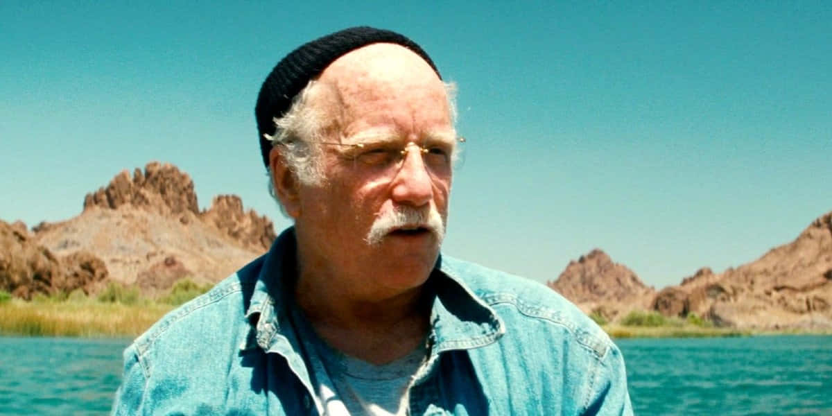 演員 桌布 壁紙 — 一位留著白色鬍鬚、戴著黑色毛帽的長者，在清澈藍天下，若有所思地凝視著被崎嶇沙漠岩層環繞的平靜湖泊。