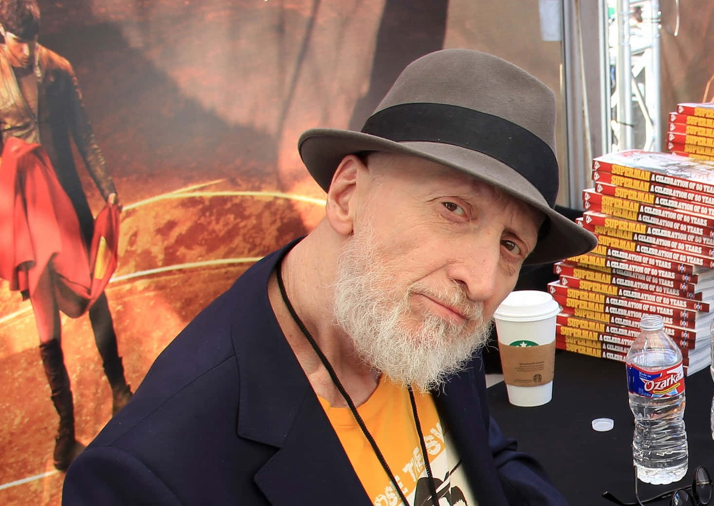 Frank Miller 桌布 壁紙 — 一位留著白鬍子、戴灰色軟呢帽的男子，在堆疊的超人書籍和星巴克杯子旁對著鏡頭微笑。