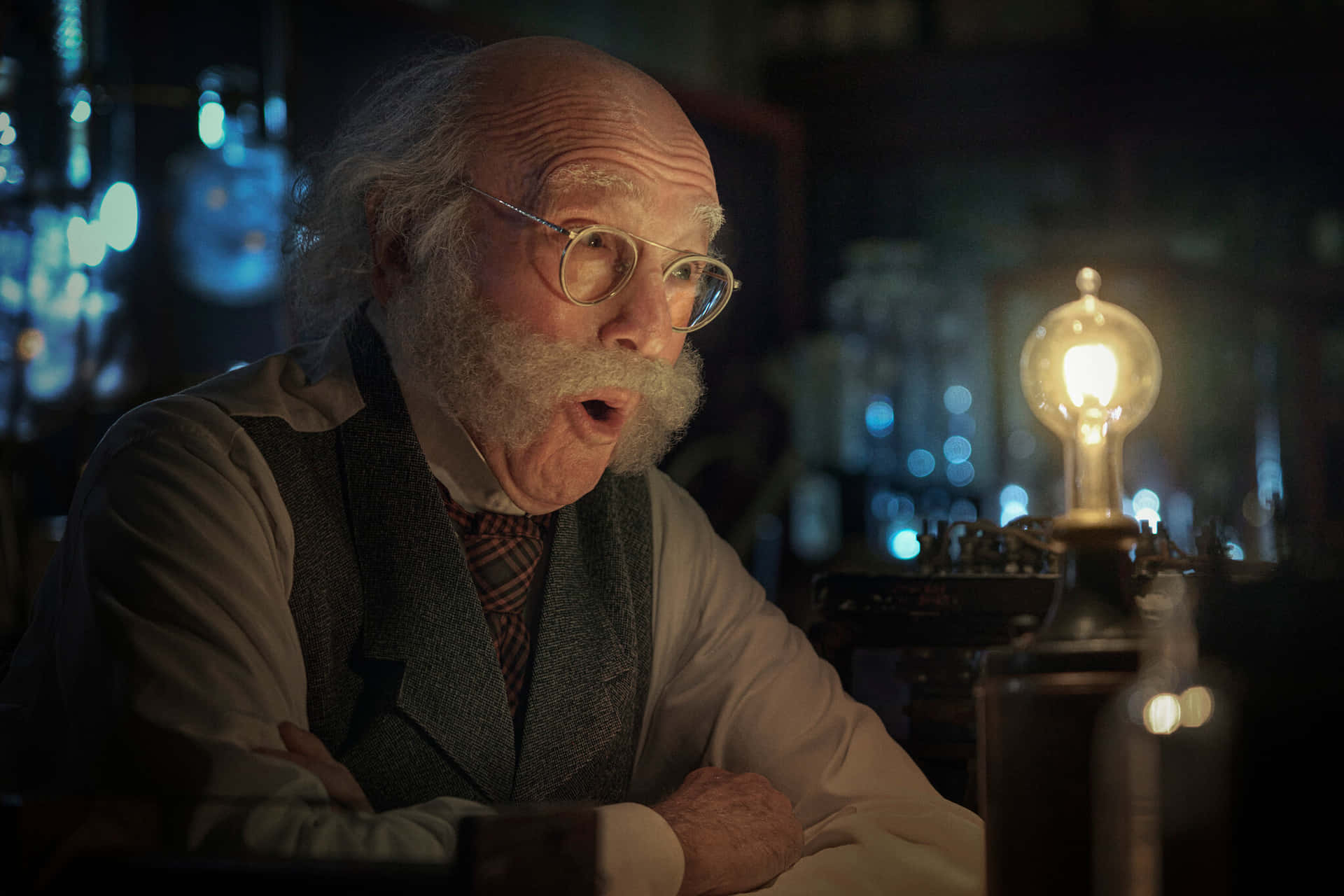 larry david 桌布 壁紙 — 一位留著白鬍子、戴著眼鏡的年長科學家，在一個裝滿玻璃瓶、光線昏暗的實驗室裡，敬畏地凝視著一個發光的燈泡。