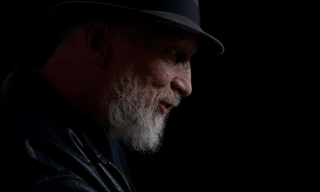 Frank Miller 桌布 壁紙 — 一位留著白鬍子、戴著軟呢帽的男子，在黑色背景前以戲劇性的側面輪廓呈現。