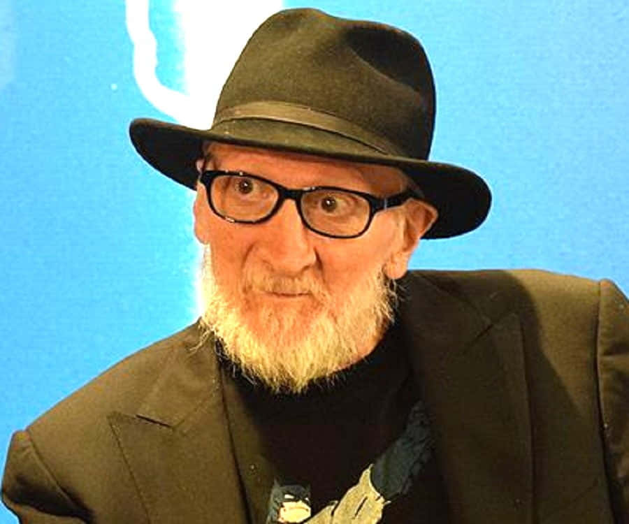 Frank Miller 桌布 壁紙 — 一位留著白鬍子、戴著黑色眼鏡的老年男子，在明亮的藍色背景前戴著黑色軟呢帽、穿著西裝。