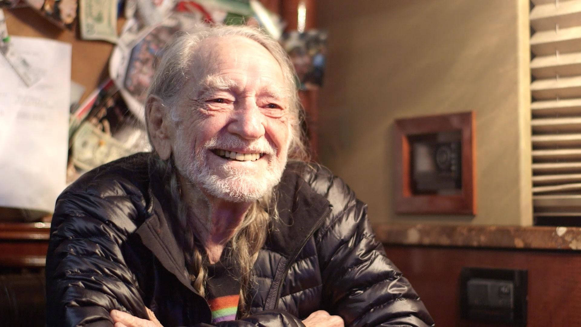 smiling 桌布 壁紙 — 一位留著白鬍子和長髮的長者，在室內穿著黑色羽絨外套，露出溫暖的笑容。