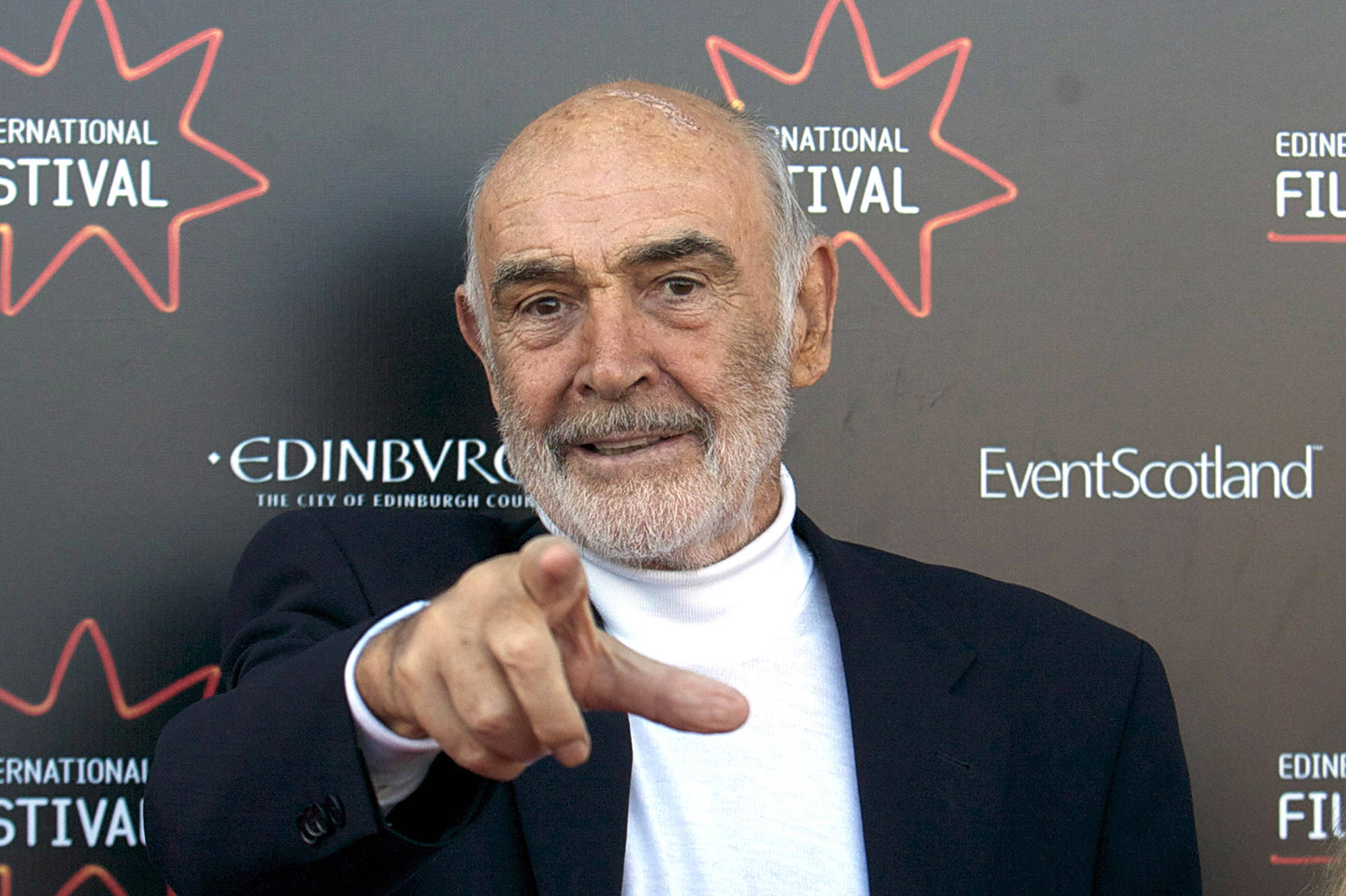 Sean Connery 桌布 壁紙 — 一位留著白鬍子的尊貴長者，在帶有橙色星星標誌和節日文字的深色背景下，直接指向相機。