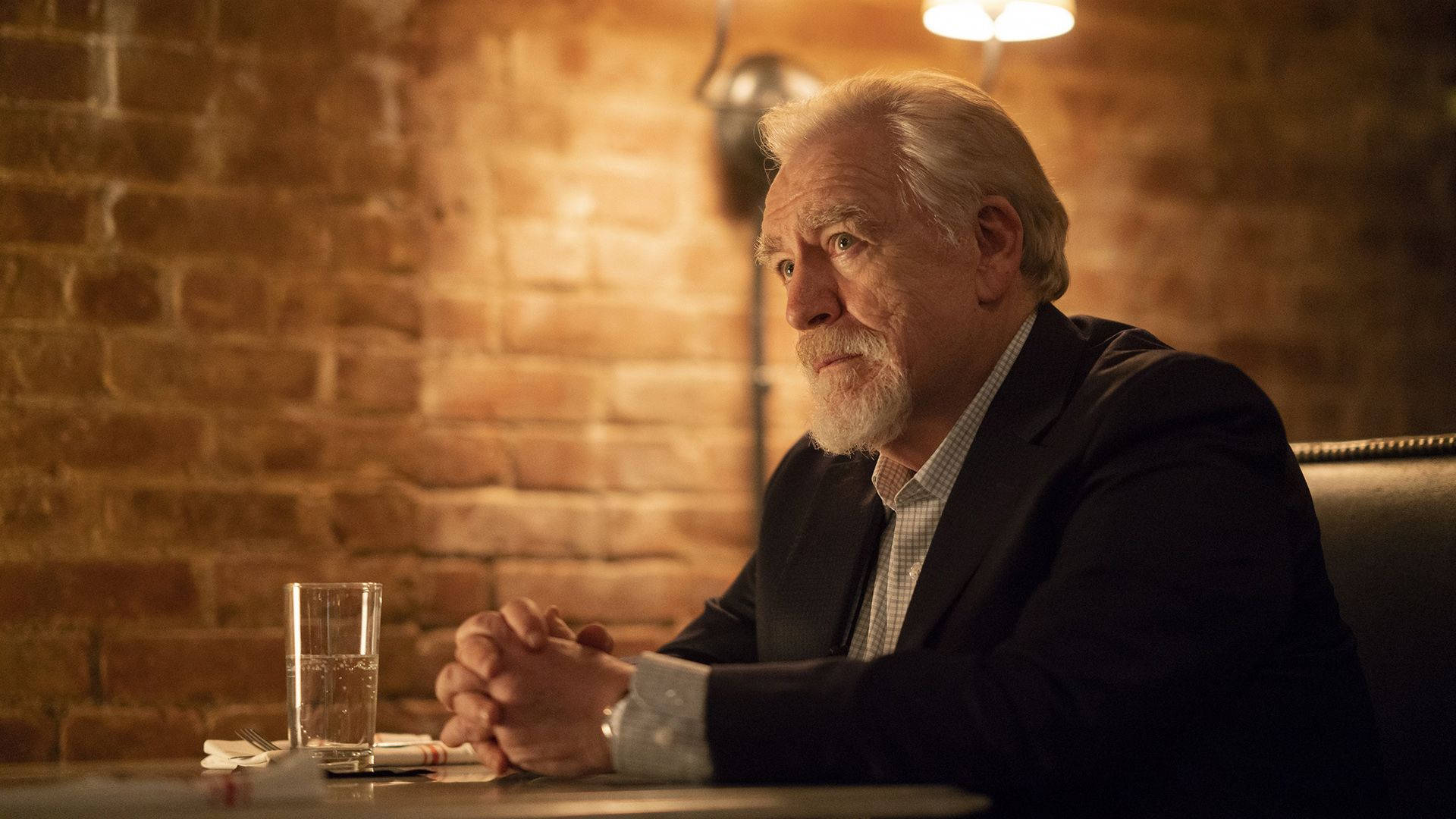 Succession 桌布 壁紙 — 一位留著白鬍子的年長男士坐在一間磚牆餐廳燈光昏暗的桌子旁，雙手緊握，若有所思地凝視遠方。
