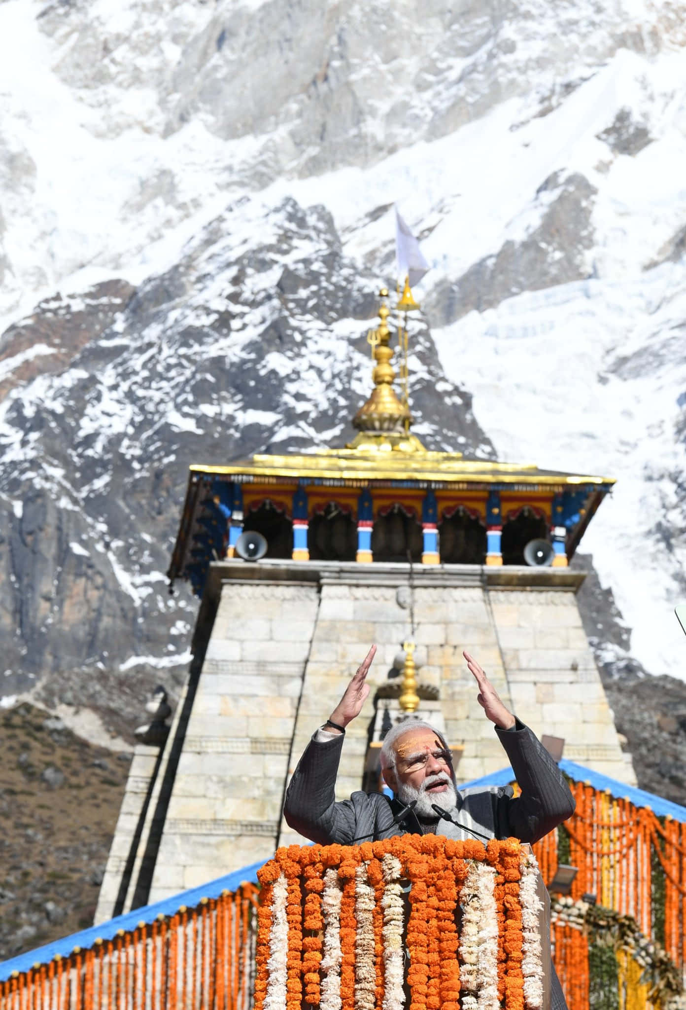 Kedarnath 桌布 壁紙 — 一位留著白鬍子的長者，在裝飾華麗的講台上，以金色佛塔為背景，背後是白雪覆蓋的喜馬拉雅山峰，熱情地演講。