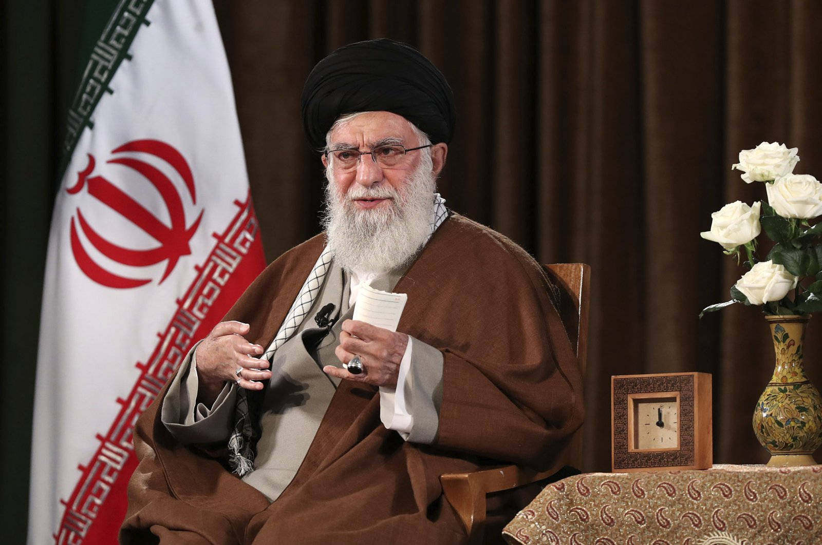 Iran 桌布 壁紙 — 一位留著白鬍鬚的年長宗教領袖坐在椅子上，手持文件，旁邊有伊朗國旗和白玫瑰。