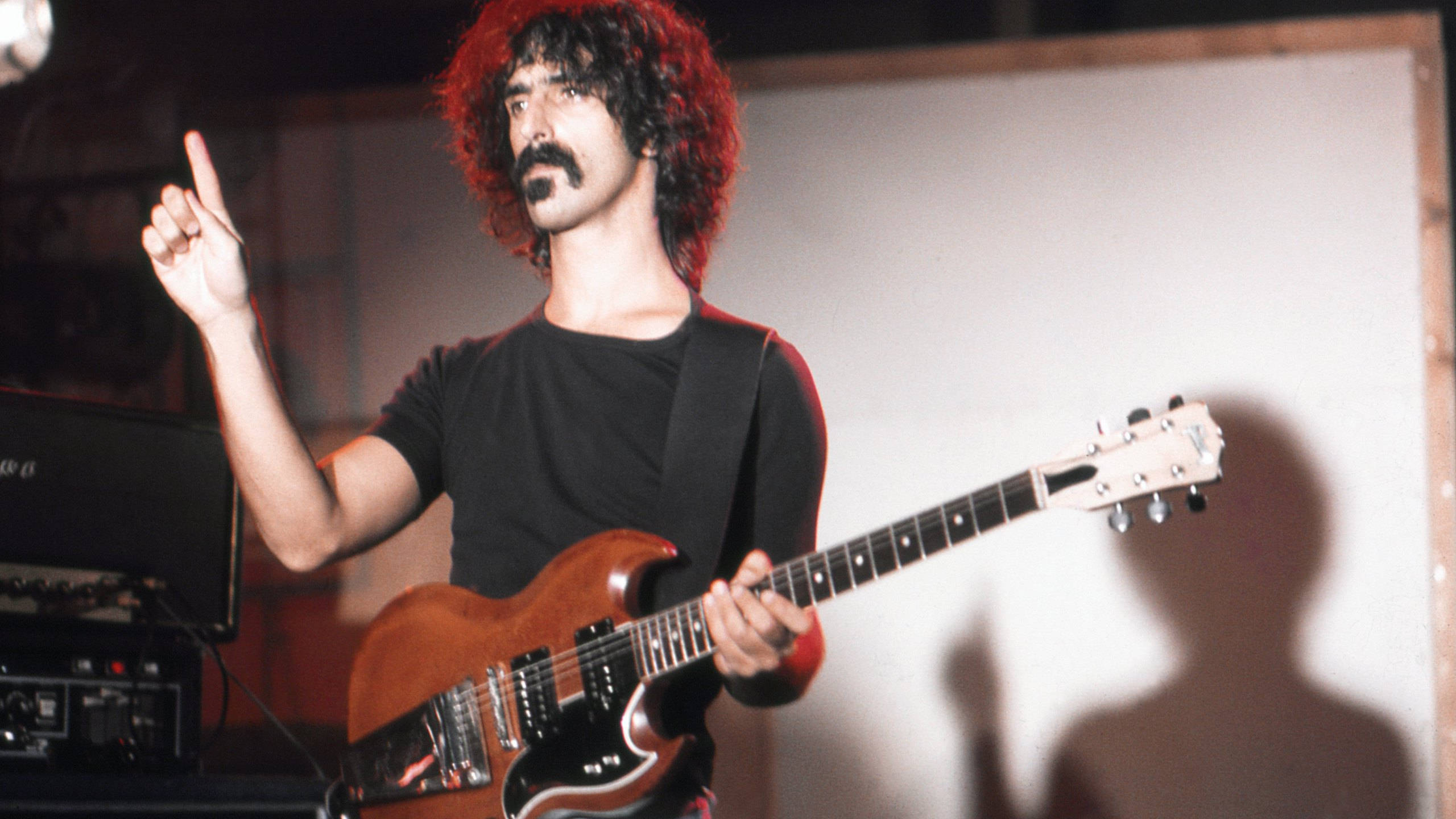 Zappa 桌布 壁紙 — 一位留著紅色捲髮和鬍子的搖滾吉他手，在舞台上彈奏一把旭日色電吉他，同時向上指著。