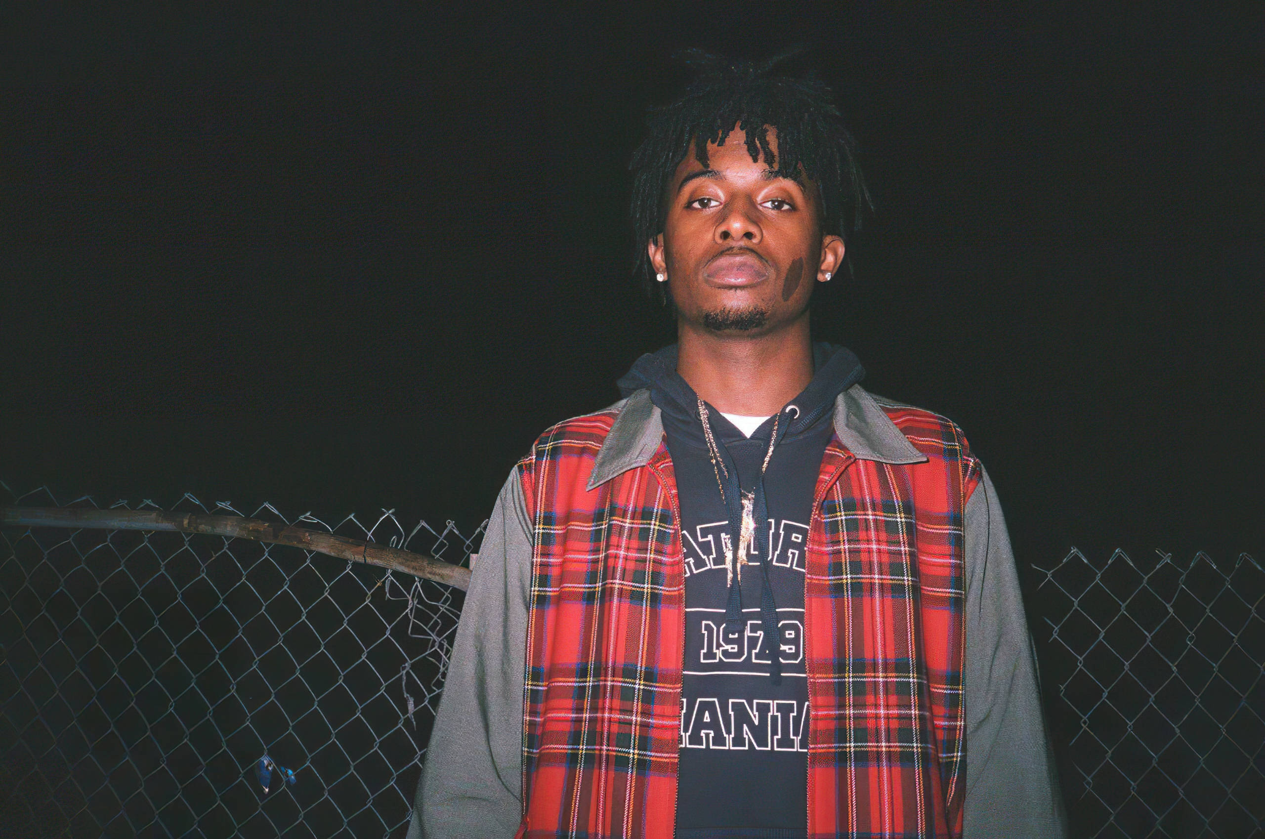 Carti 桌布 壁紙 — 一位留著辮子的年輕男子在夜晚自信地站在鐵絲網圍欄前，身穿紅色格紋背心配連帽上衣。