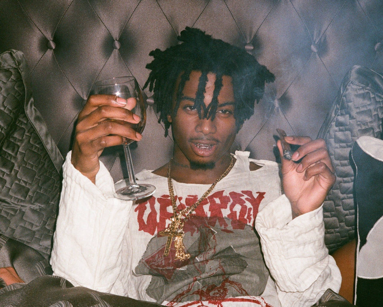 Carti 桌布 壁紙 — 一位留著辮子的年輕男子坐在簇絨皮革沙發上，手持酒杯，抽著大麻煙，看著鏡頭。
