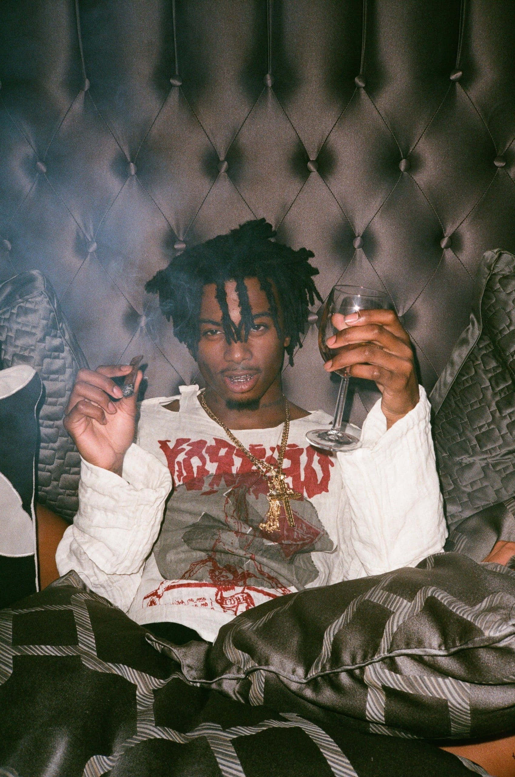 playboi carti pfp 桌布 壁紙 — 一位留著辮子的年輕男子懶洋洋地躺在簇絨皮革沙發上，手持酒杯，在煙霧瀰漫的昏暗燈光下抽煙。