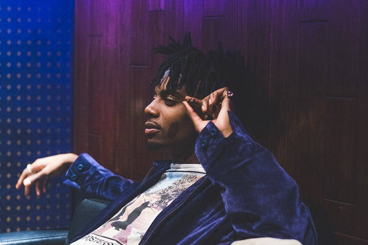 Carti 桌布 壁紙 — 一位留著辮子的年輕男子穿著天鵝絨外套，靠著木鑲板牆壁，沐浴在紫色和藍色燈光中。