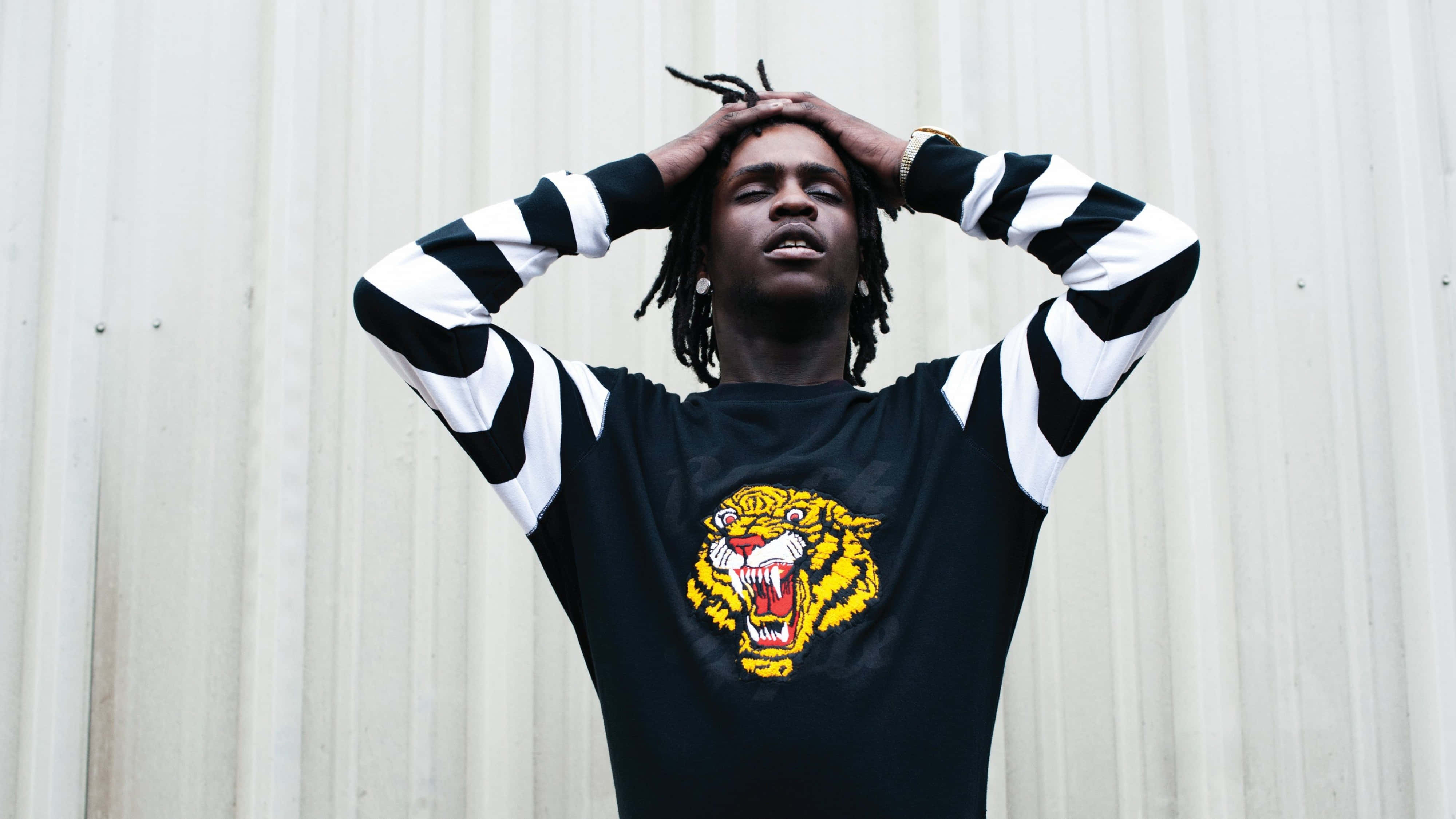 4K Rapper 桌布 壁紙 — 一位留著辮子的年輕男子穿著黑白條紋襯衫，上面有黃色老虎圖案，頭靠著波紋牆。