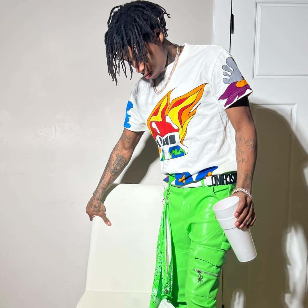 Youngboy 桌布 壁紙 — 一位留著辮子的年輕男子靠在一張白色椅子上，穿著色彩繽紛的圖案T恤和螢光綠色褲子，手持一個白色杯子。