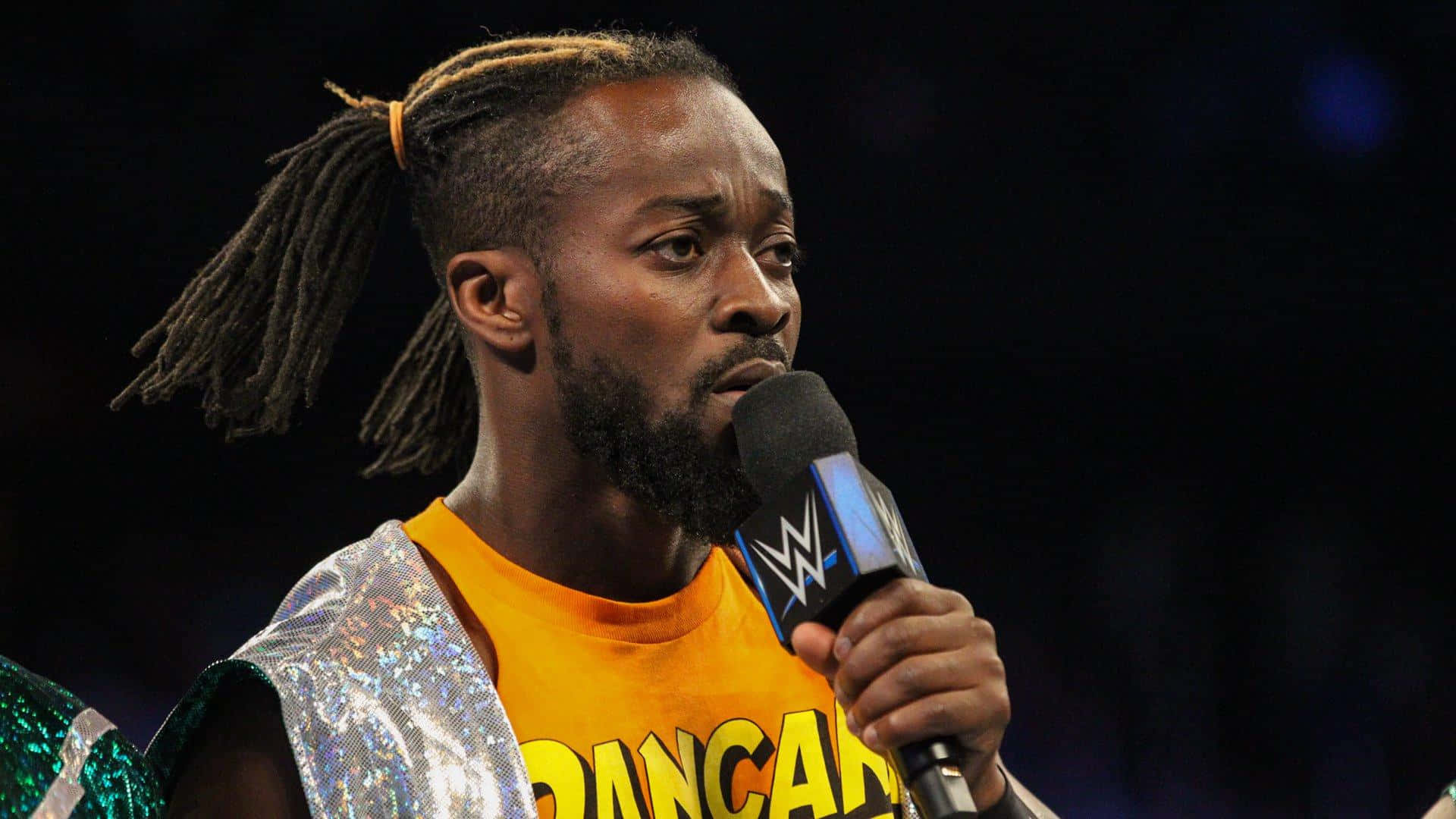 WWE 桌布 壁紙 — 一位留著辮子的摔角手，身穿閃亮的銀綠色背心，內搭橙色襯衫，對著WWE麥克風說話。