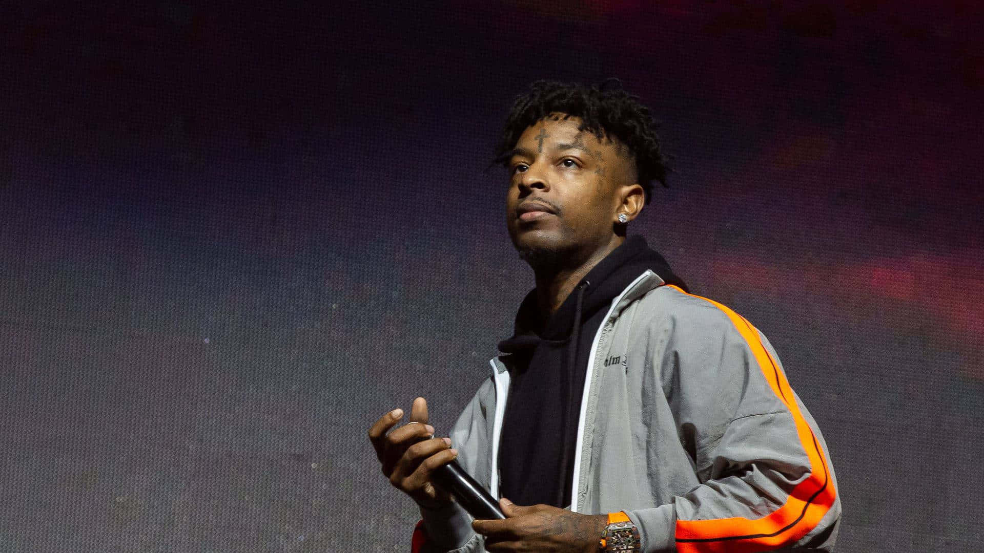 21 Savage 桌布 壁紙 — 一位留著辮子的男性表演者在舞台上，在戲劇性的紫色和紅色燈光下握著麥克風。
