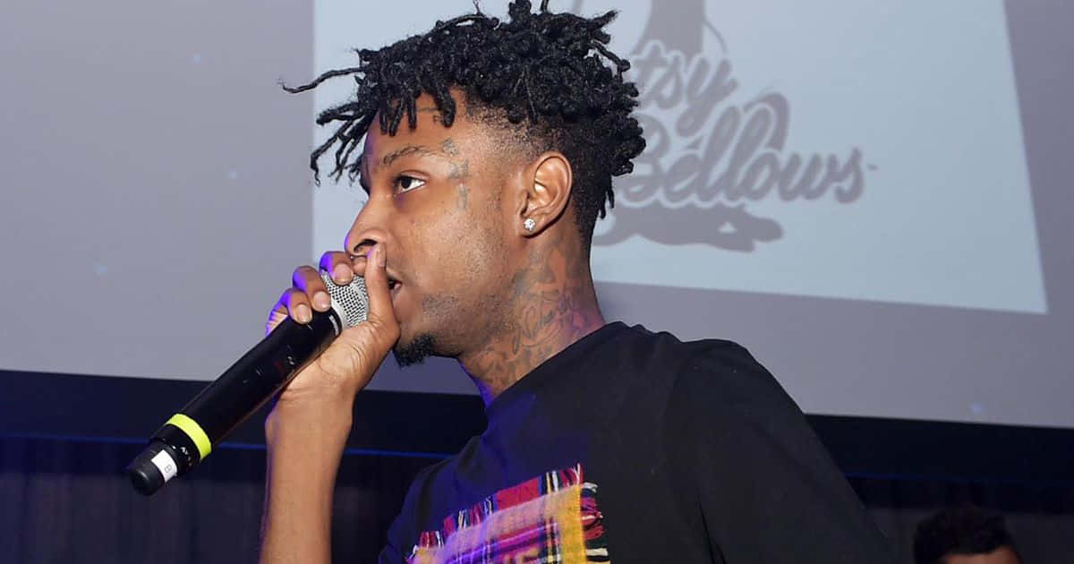 21 Savage 桌布 壁紙 — 一位留著辮子的男饒舌歌手在紫色燈光下於舞台上熱情地對著麥克風表演。