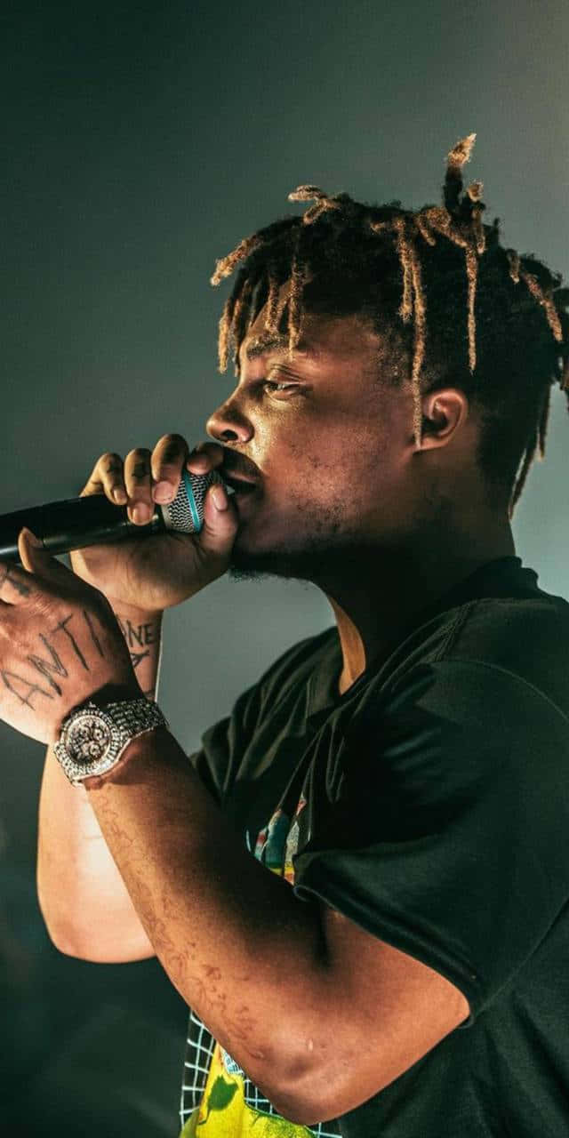Juice Wrld iPhone 桌布 壁紙 — 一位留著辮子的男饒舌歌手在舞台燈光下對著麥克風唱歌，身穿黑色襯衫，戴著鑲鑽手錶。