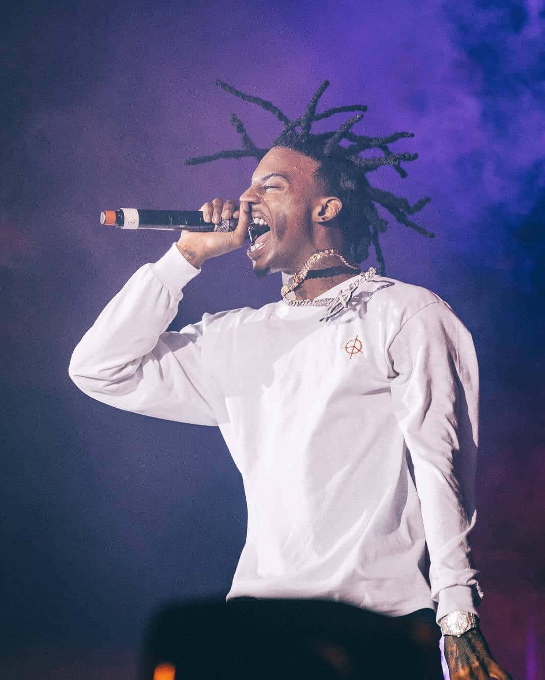 Playboi Carti HD 桌布 壁紙 — 一位留著辮子的說唱歌手在紫色舞台燈光下熱情地對著麥克風唱歌。