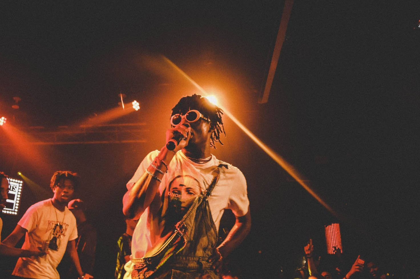 Carti 桌布 壁紙 — 一位留著辮子頭、戴圓框眼鏡的饒舌歌手，在昏暗的演唱會場地中，於溫暖的舞台燈光下熱情地對著麥克風表演。