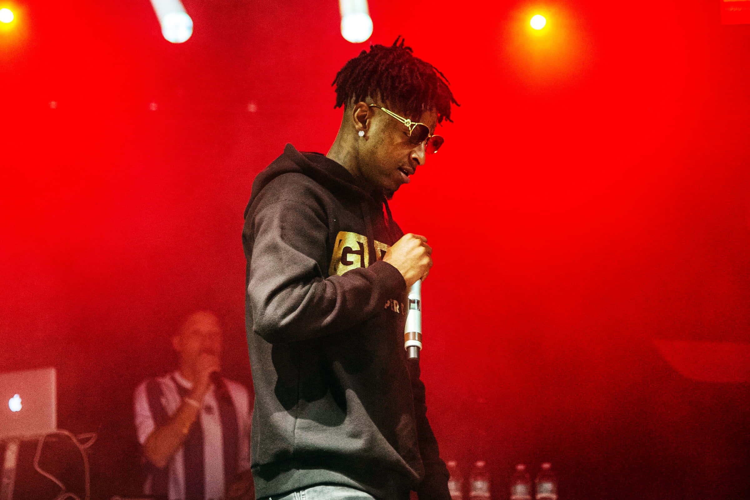 21 Savage 桌布 壁紙 — 一位留著辮子頭、戴金邊眼鏡的饒舌歌手在強烈紅光下拿著麥克風在舞台上表演。