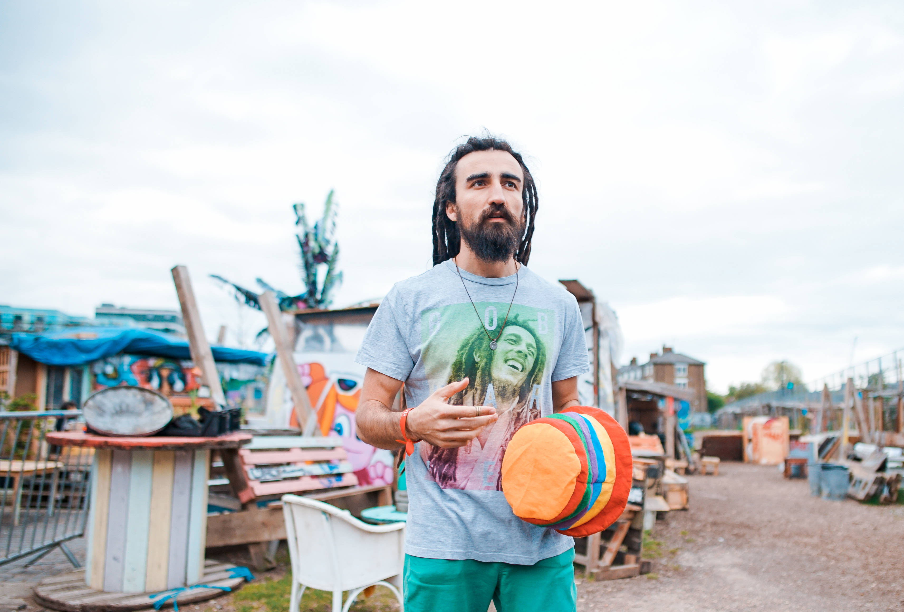 Reggae 桌布 壁紙 — 一位留著辮子頭、鬍鬚濃密的男士站在雜亂的戶外垃圾場，手持彩色飛盤，身穿Bob Marley T恤。