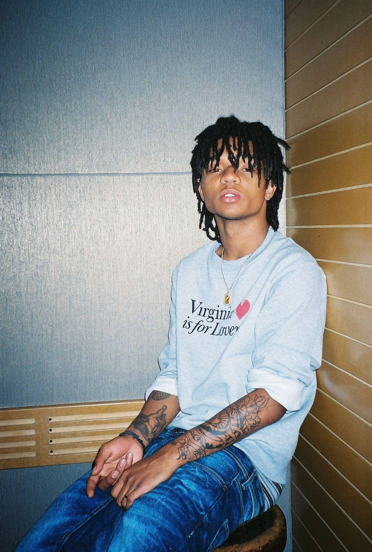 Swae Lee 桌布 壁紙 — 一位留著辮子頭和刺青的年輕男子，隨意地坐在木長椅上，背靠有紋理的牆壁，身穿維吉尼亞運動衫。
