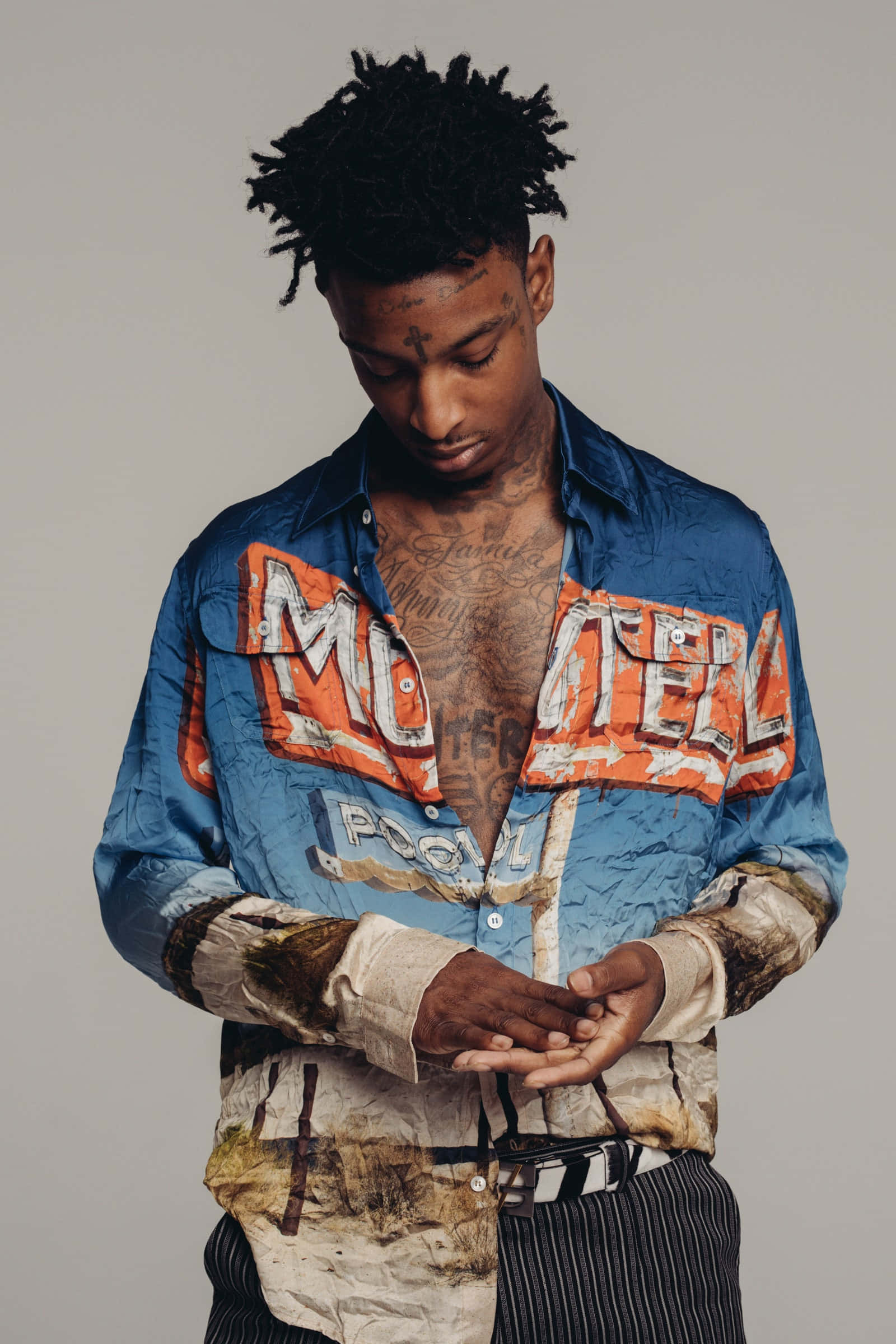 21 Savage 桌布 壁紙 — 一位留著辮子頭和刺青的年輕男子，穿著一件帶有橙色字樣的破舊藍色襯衫，低頭看著自己的手。