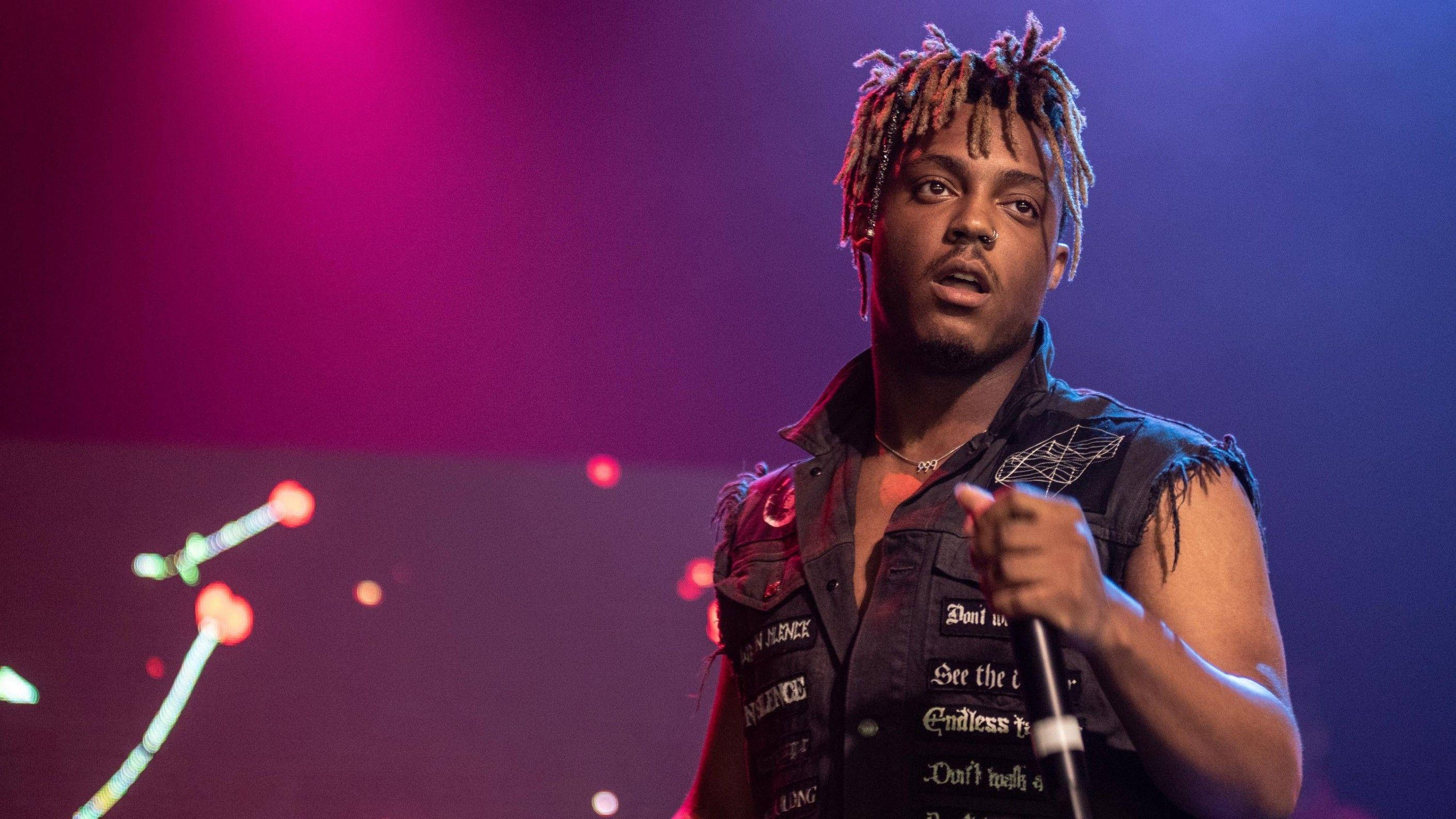 Juice Wrld Desktop 桌布 壁紙 — 一位留著辮子頭的男表演者在充滿活力的粉紅色和藍色舞台燈光下對著麥克風唱歌。
