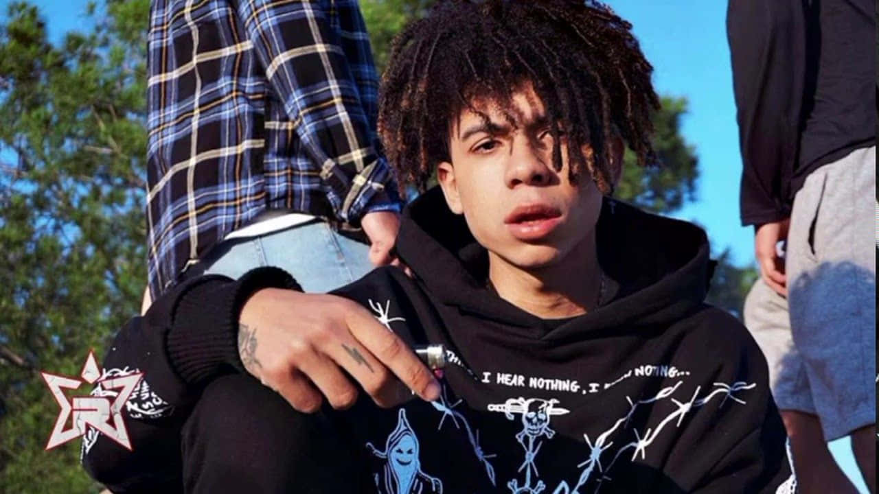 Hip Hop 桌布 壁紙 — 一位留著辮子頭的青年男子，手持筆，眼神專注，身穿帶有藍色圖案印花的黑色連帽衫，在戶外明亮的藍天下，附近有樹木和朋友。