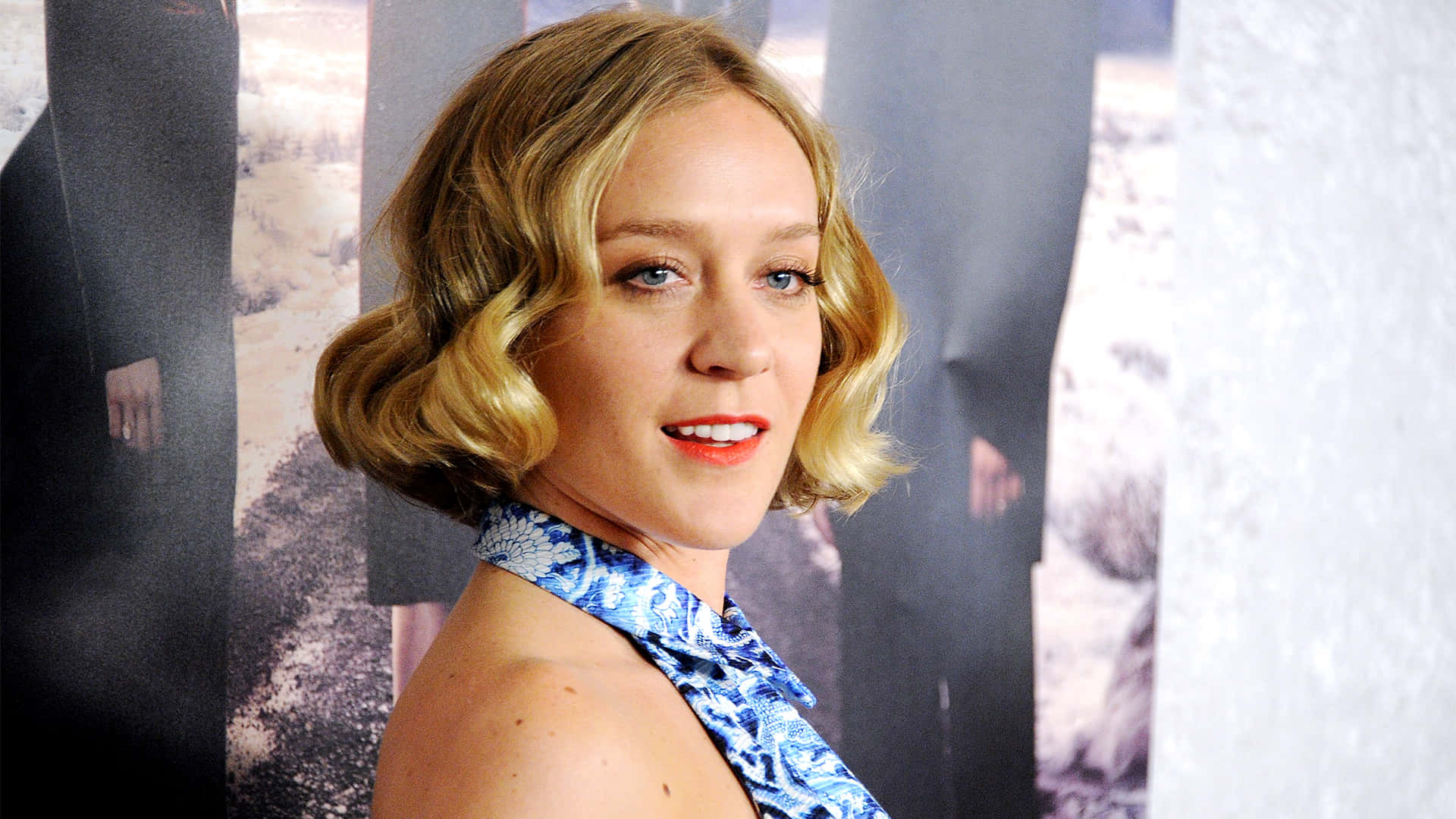 Chloë Sevigny 桌布 壁紙 — 一位留著金色波浪短髮、塗著紅色唇膏的女性，微笑著穿著藍色圖案掛頸上衣。