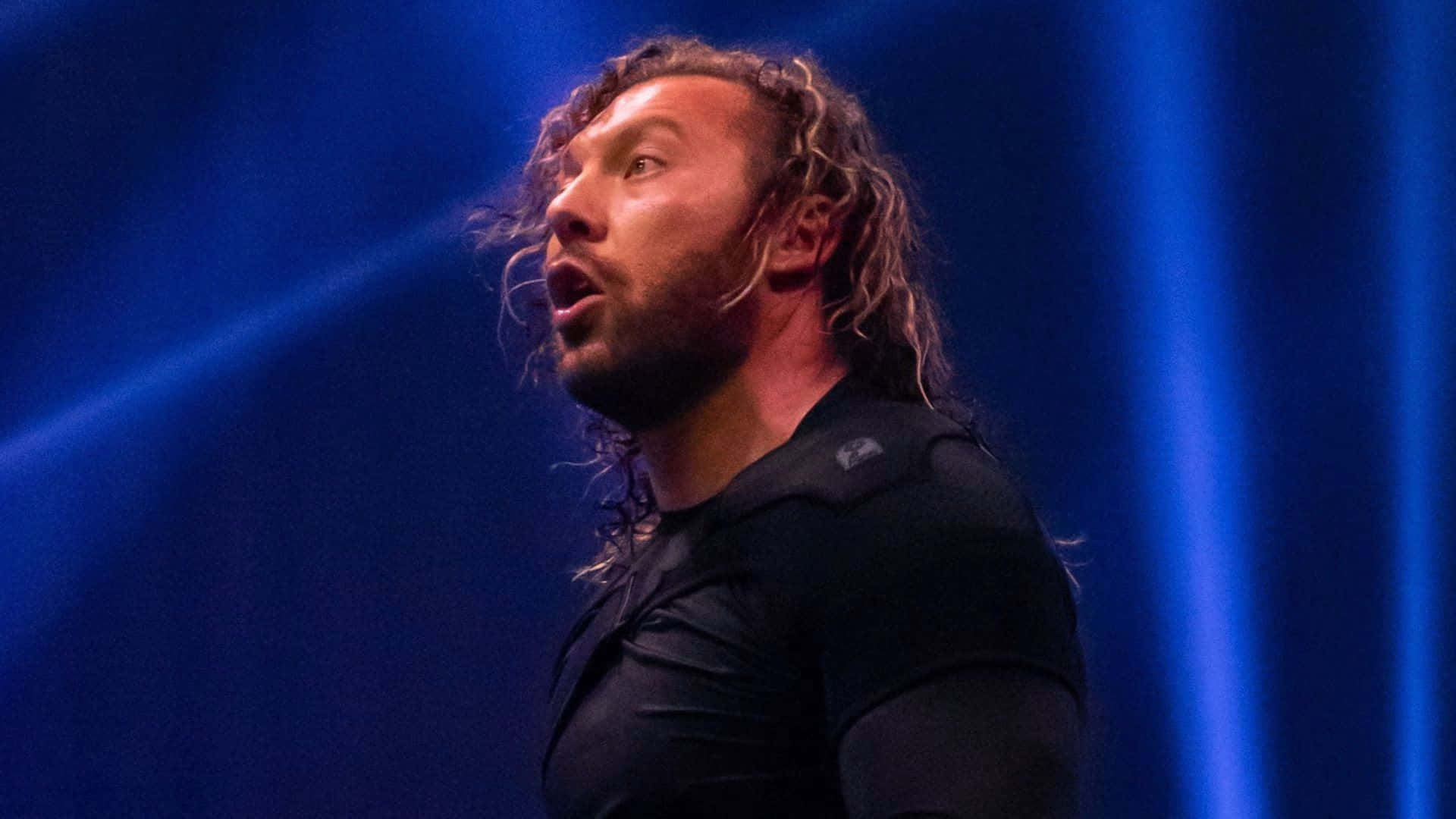 Aew 桌布 壁紙 — 一位留著長捲髮和鬍鬚的摔角手在戲劇性的藍色舞台燈光下咆哮。