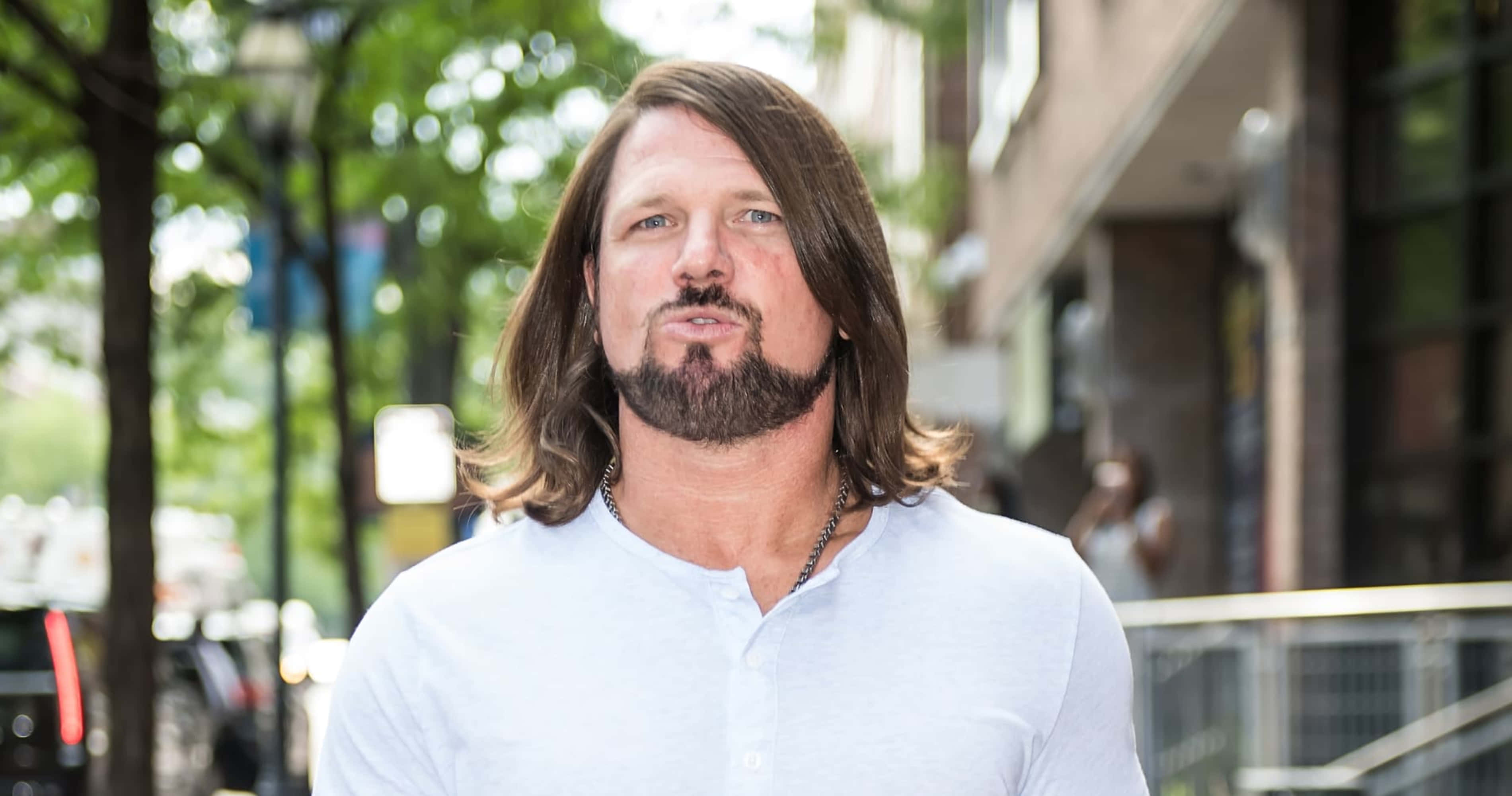 WWE 桌布 壁紙 — 一位留著長棕色頭髮和鬍鬚的男人自信地站在陽光普照的城市人行道上，周圍環繞著樹木和建築物。