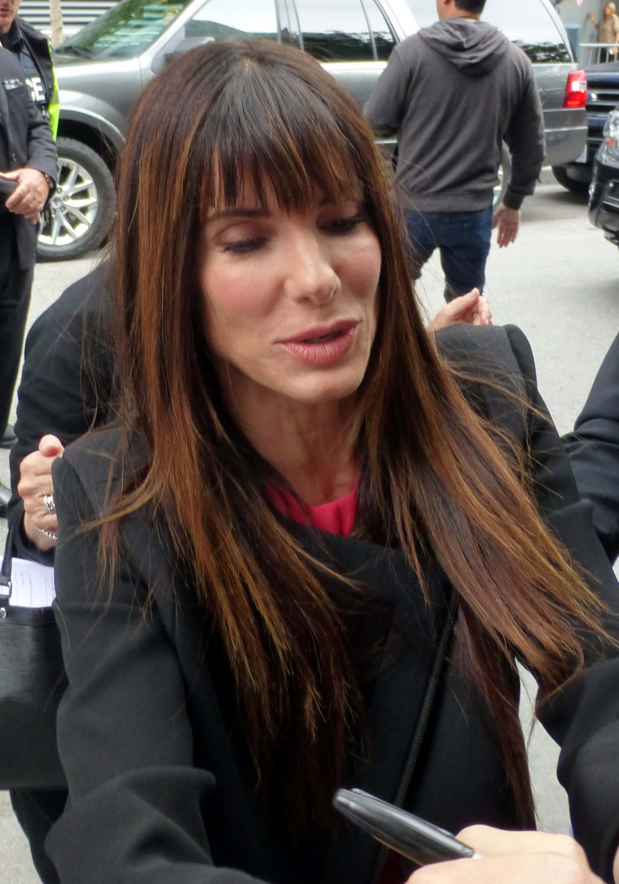 Sandra Bullock 2021 桌布 壁紙 — 一位留著長棕髮和瀏海的女人，在城市的街道上被汽車和人群包圍著簽名。