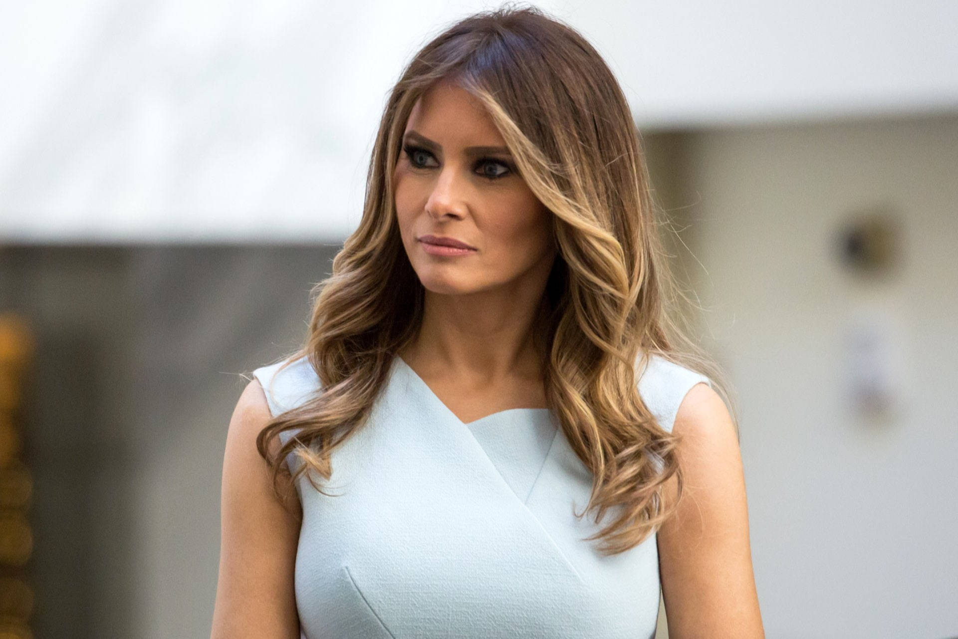 Melania 桌布 壁紙 — 一位留著長波浪棕色頭髮、身穿淺藍色無袖連衣裙的女性，表情嚴肅地看向一旁。