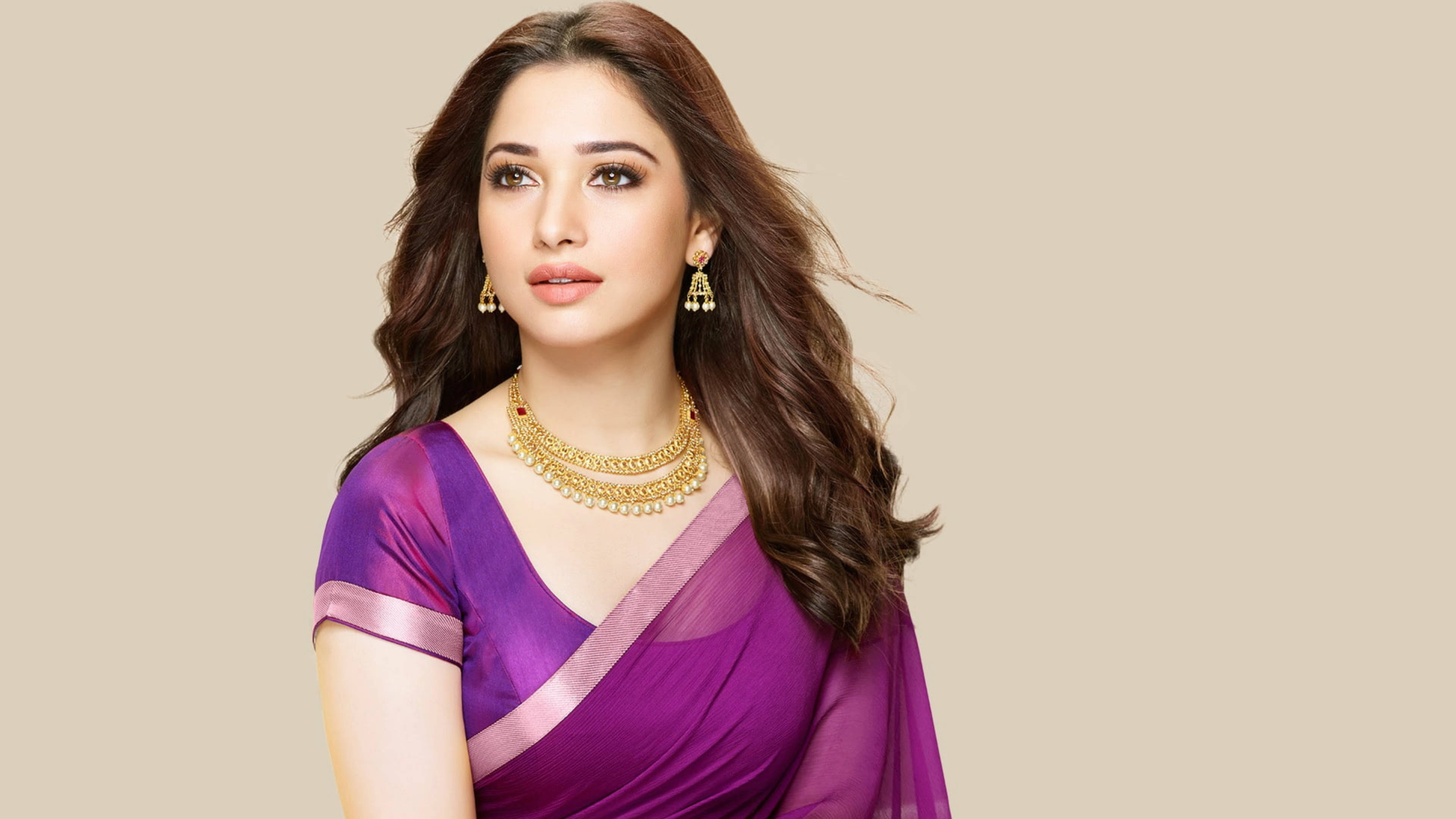 tamannaah bhatia 桌布 壁紙 — 一位留著長波浪棕色頭髮的女性，在米色中性背景前穿著鮮豔的紫色紗麗，佩戴金色珠寶。