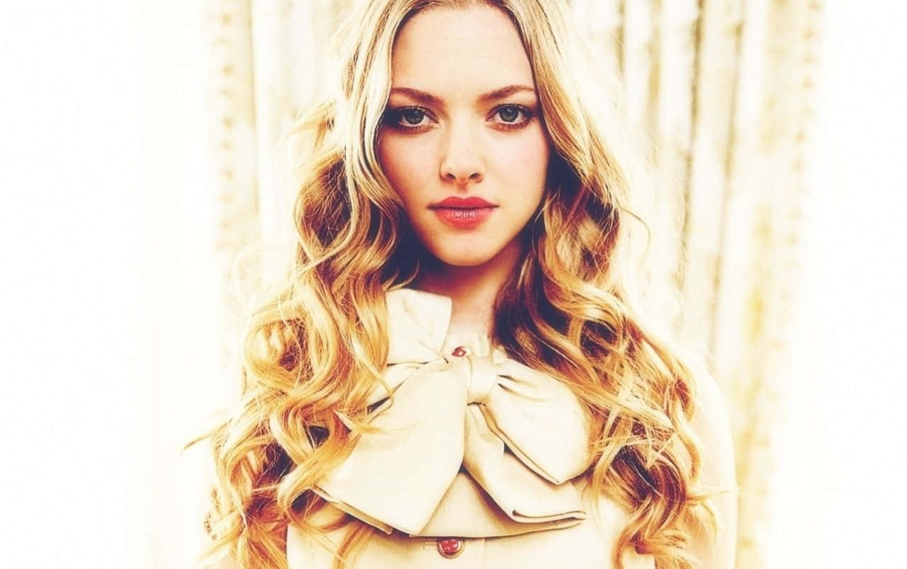 Amanda Seyfried 桌布 壁紙 — 一位留著長波浪金髮的女性，在柔和模糊的背景前穿著一件飾有蝴蝶結的米色外套。