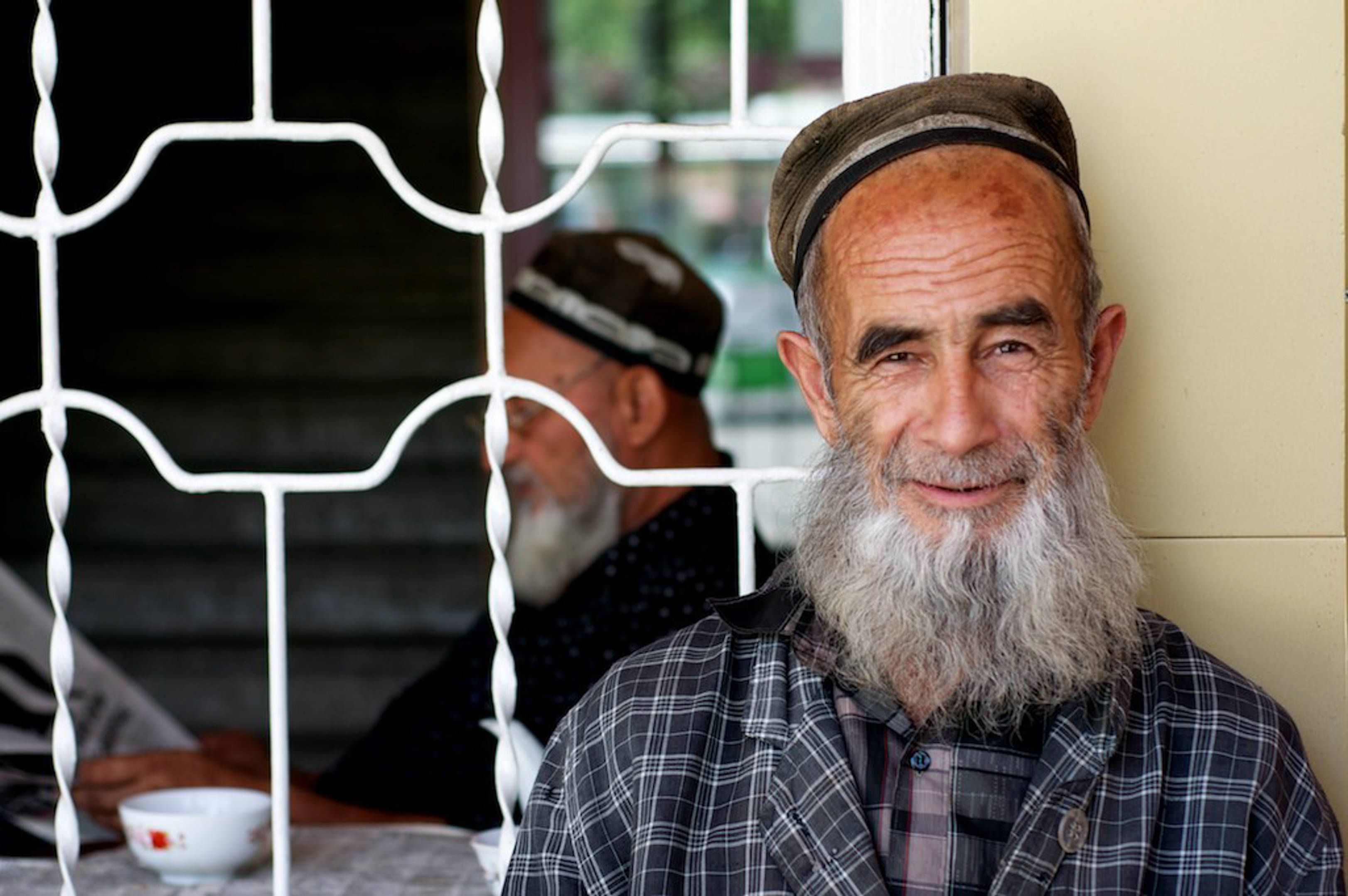 Tajikistan 桌布 壁紙 — 一位留著長灰鬍鬚、戴著傳統帽子的長者，在白色裝飾窗格後面溫暖地微笑。
