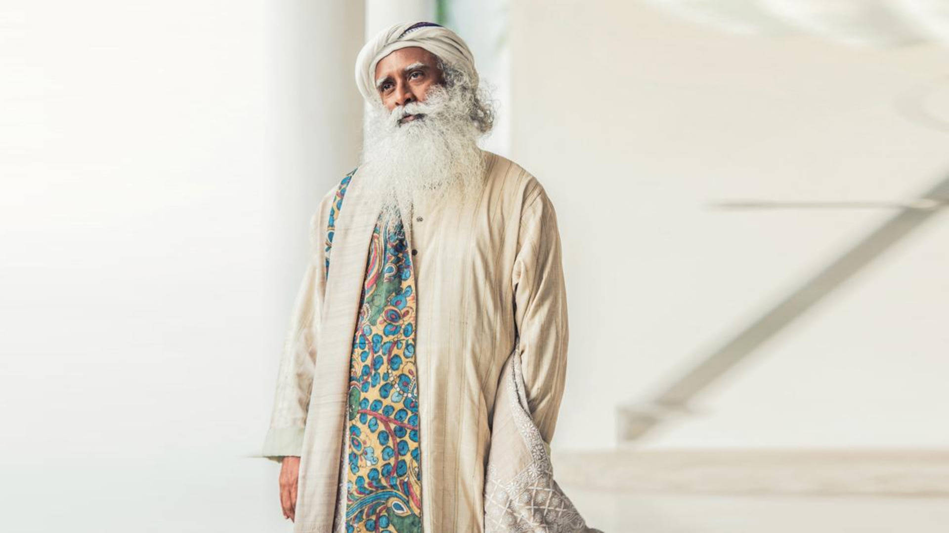 Indian 桌布 壁紙 — 一位留著長白鬍子、戴著頭巾的長者，身著圖案長袍，站在明亮的簡約室內。