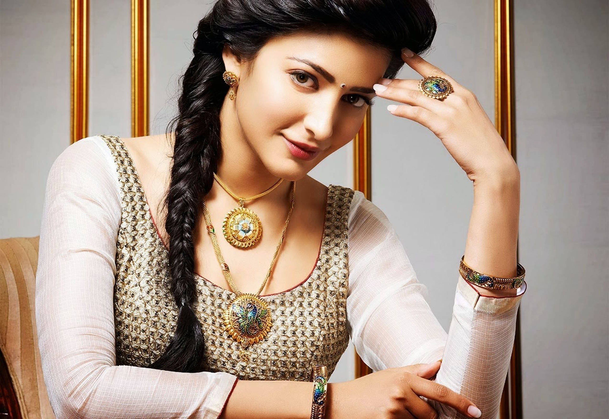 Shruti Hassan HD 桌布 壁紙 — 一位留著長辮子、佩戴華麗珠寶的女性，在金色邊框的背景前優雅地擺姿勢。