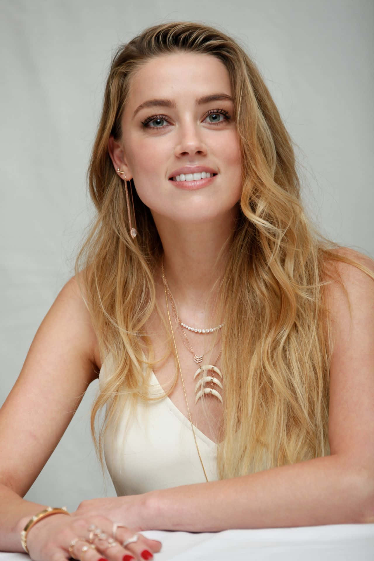 Amber Heard 桌布 壁紙 — 一位留著長金髮的女性，戴著多層金飾和米白色上衣，坐在桌旁溫暖地微笑。
