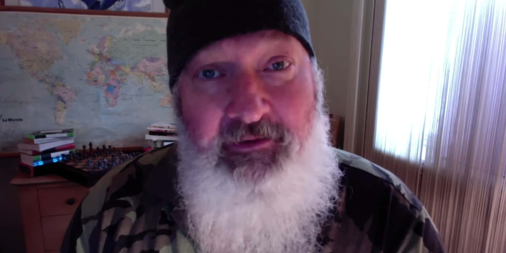 Randy Quaid 桌布 壁紙 — 一位留著長長白鬍子、戴著毛帽的男人，在一個有世界地圖和棋盤的房間裡直視鏡頭。