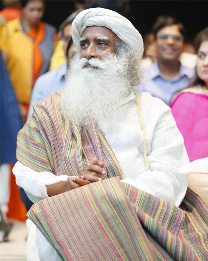 Sadhguru 桌布 壁紙 — 一位留著長長白鬍子、戴著頭巾的長者，披著色彩繽紛的條紋披肩，平靜地坐在人群中。