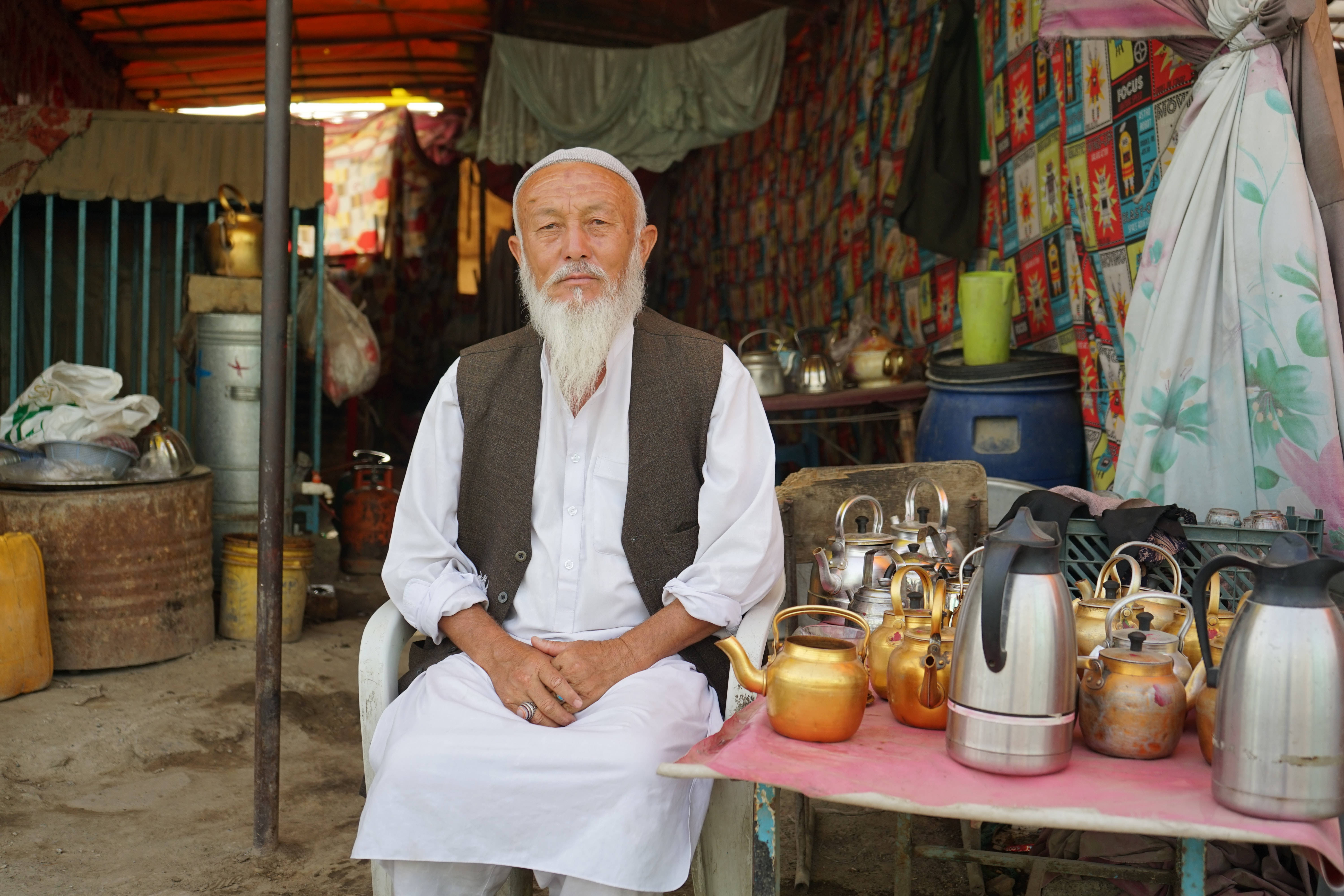 Kabul 桌布 壁紙 — 一位留著長長白鬍子的老人，平靜地坐在熙熙攘攘的市場攤位中，周圍是閃亮的黃銅和不鏽鋼水壺。
