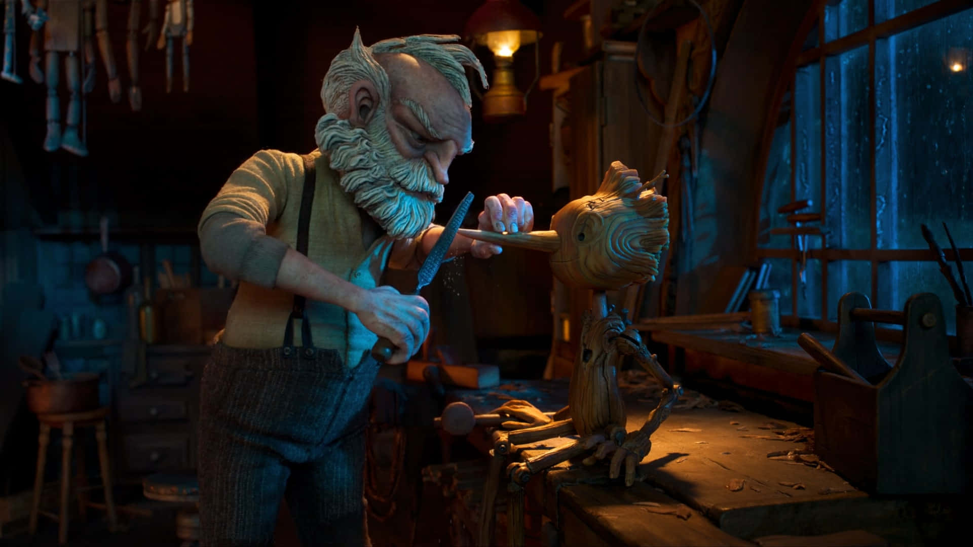 Guillermo Del Toro's Pinocchio 桌布 壁紙 — 一位留著長長白鬍子的老木雕師傅，在燈光昏暗、雜亂的工作室裡細心地雕刻著一個木偶。