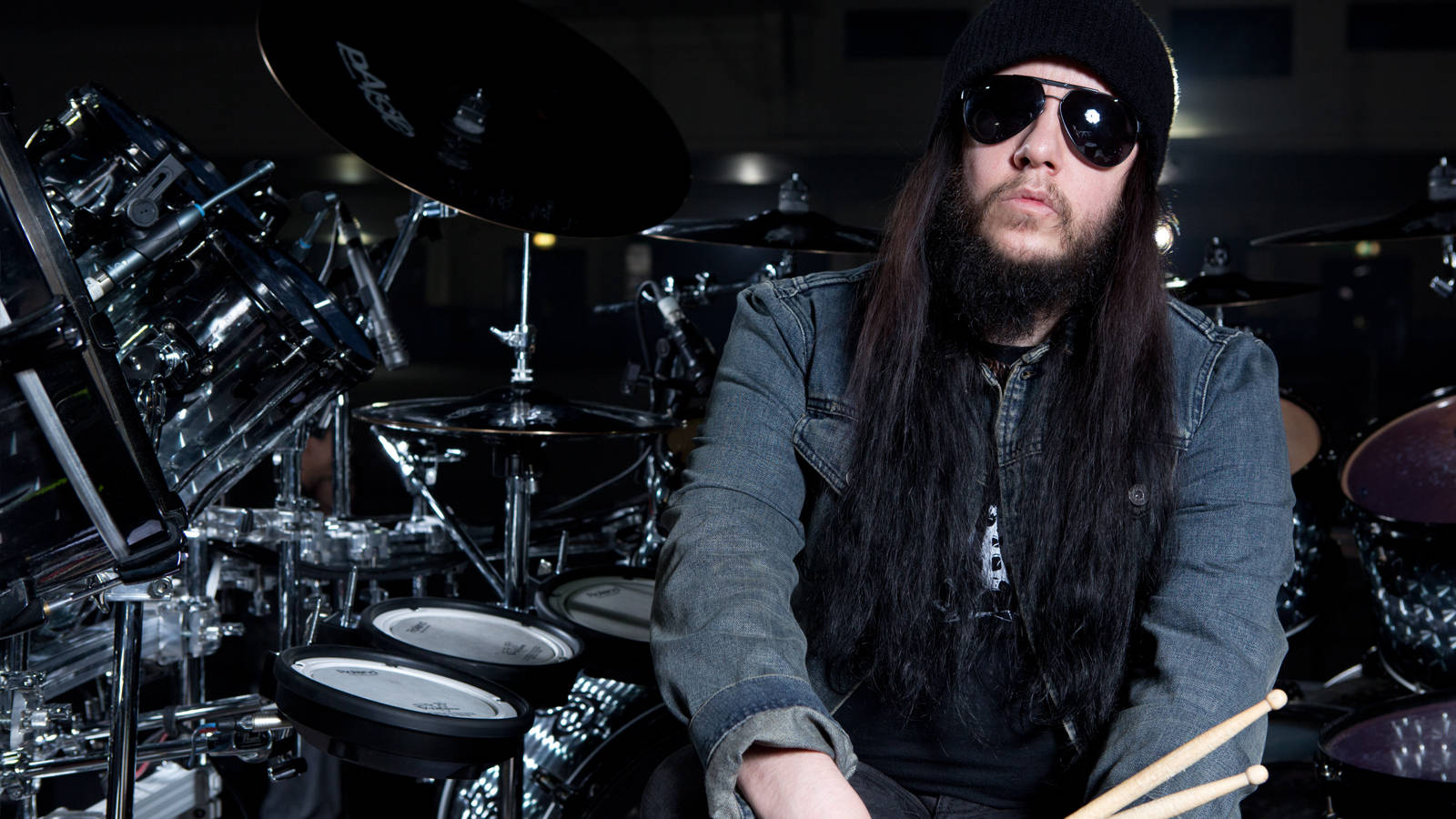 Joey Jordison 桌布 壁紙 — 一位留著長黑髮、戴著太陽眼鏡的鬍鬚鼓手，自信地坐在昏暗工作室中閃亮的chrome鼓組前。