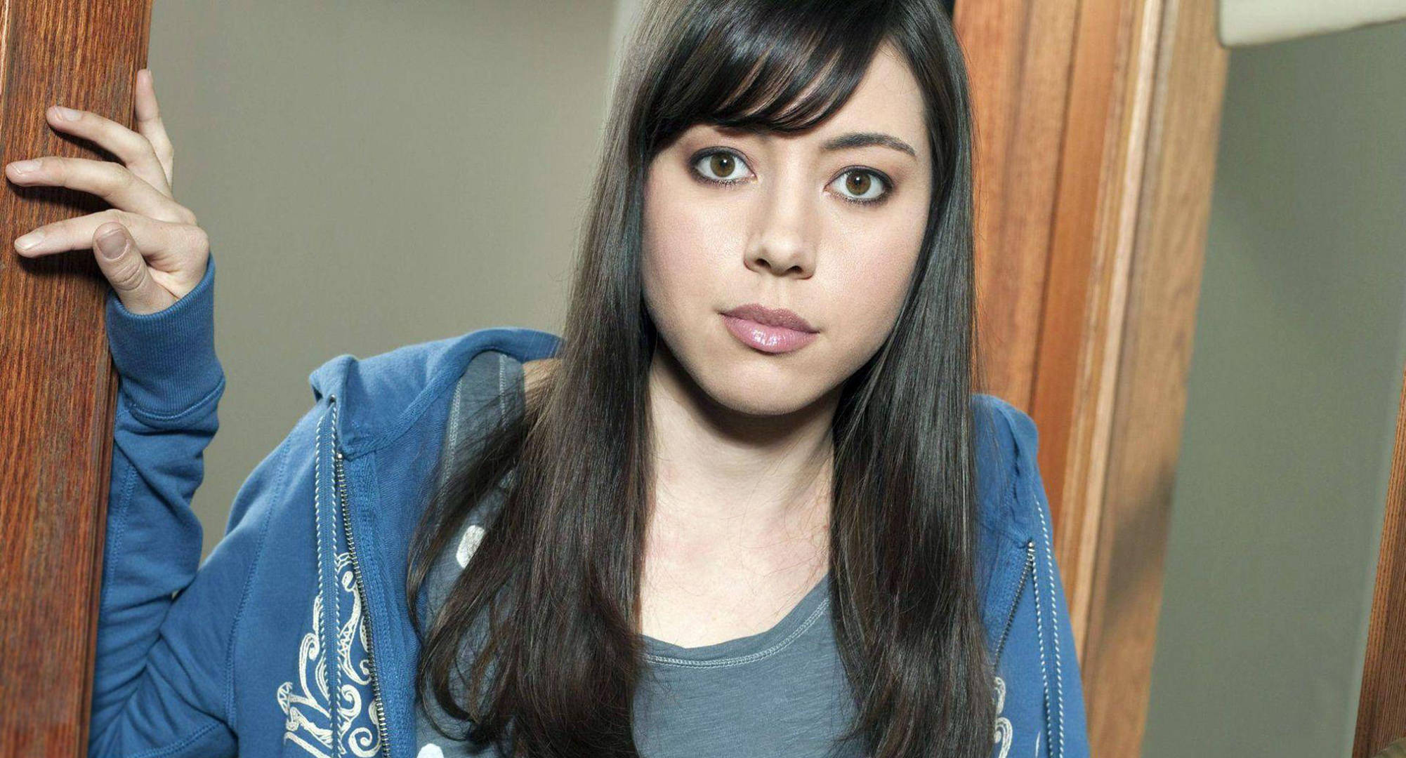 Aubrey 桌布 壁紙 — 一位留著長黑髮和瀏海的年輕女性穿著藍色連帽衫，靠著木門框直視鏡頭。