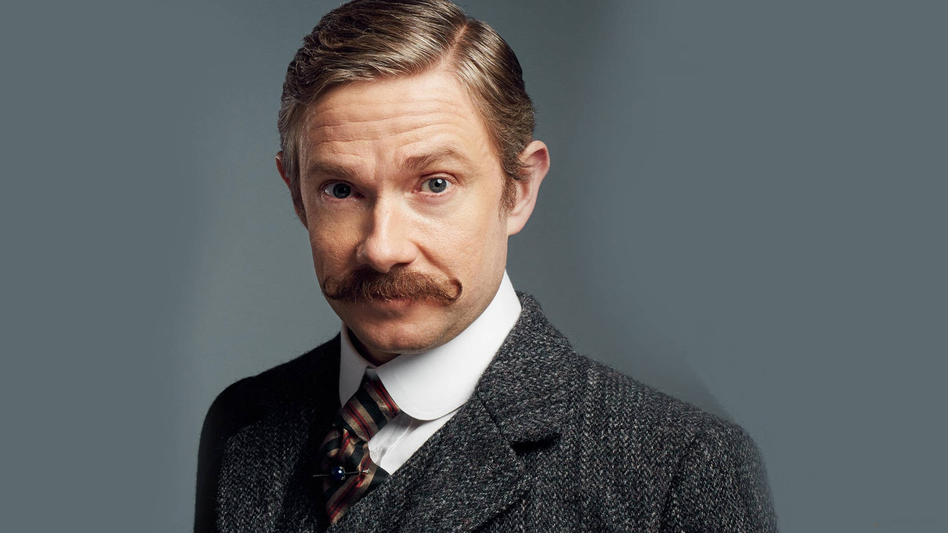Martin Freeman 桌布 壁紙 — 一位留著顯眼鬍鬚、頭髮整齊梳理的男士，穿著粗花呢西裝和圖案領帶，背景為柔和的灰色。