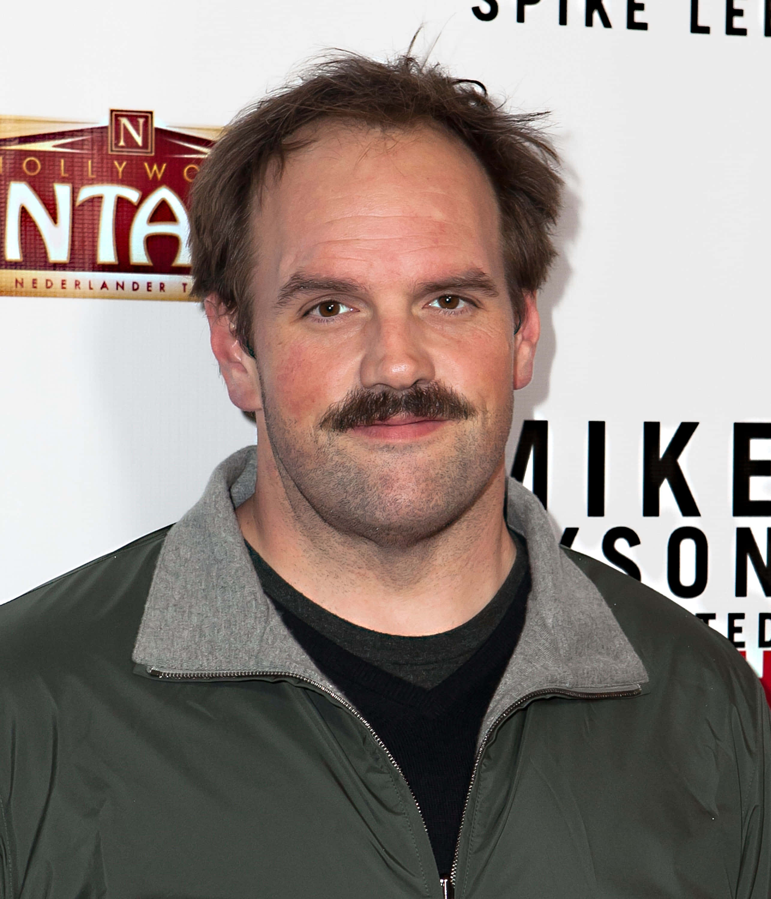 Ethan Suplee 桌布 壁紙 — 一位留著顯眼鬍鬚和凌亂頭髮的男子，在好萊塢活動中穿著綠色外套對著鏡頭微笑。