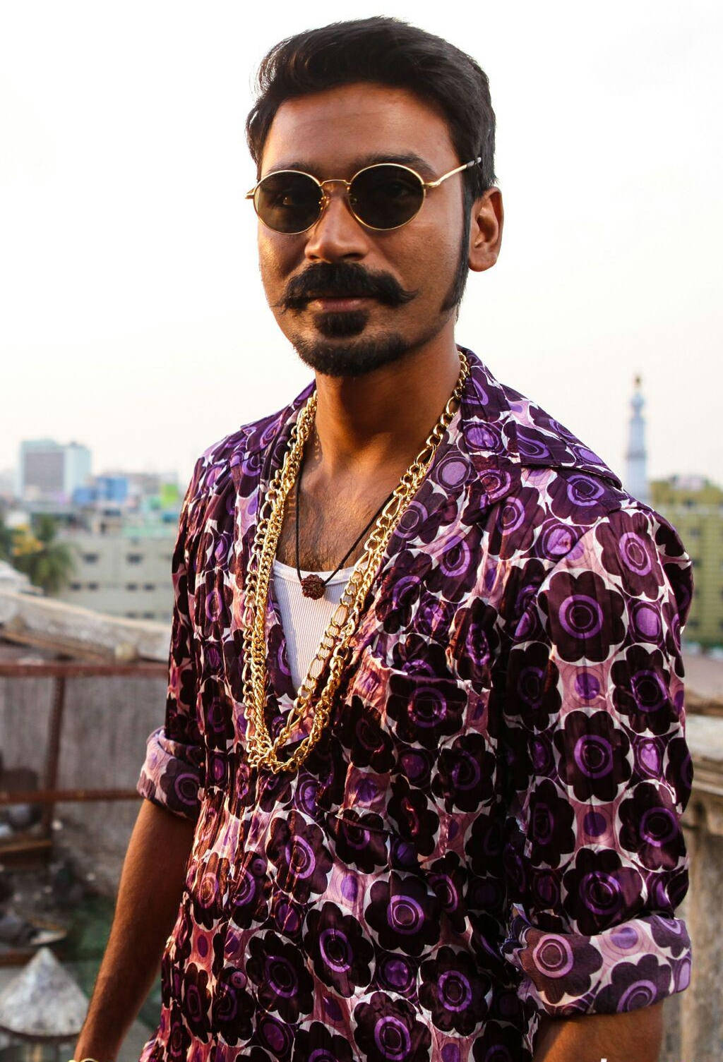 Indian 桌布 壁紙 — 一位留著鬍子、戴著金框太陽眼鏡的時尚男士，穿著紫色花卉襯衫和層疊金鍊，背景是城市屋頂。