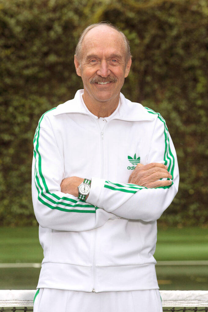 復古 桌布 壁紙 — 一位留著鬍子的年長男性，穿著白色和綠色的Adidas運動服，自信地微笑站在網球場上。